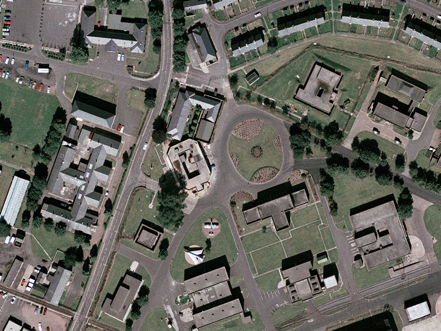 A historic colour vertical aerial photo of numerous grey-roofed buildings interspersed with roads, trees and areas of grass. The octagonal guardhouse has a flat roof and is just left of centre.