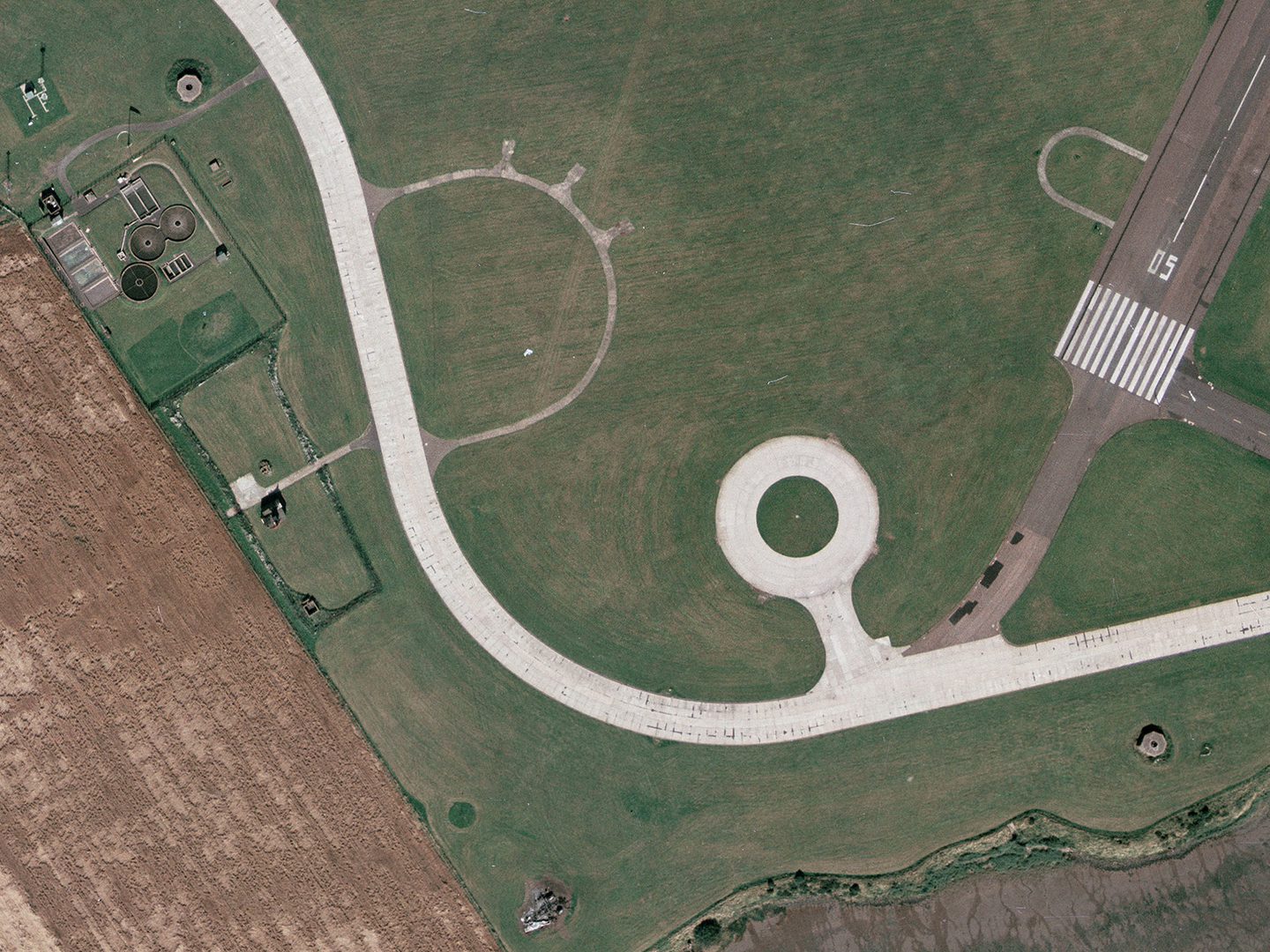 A historic colour vertical aerial photo of a light-coloured concrete road snaking through grass from the top left corner to the lower right side. 2 octagonal concrete pillboxes are visible in the top and left and bottom right of the image. Just right of centre is a circular ring of road with a grassy middle.