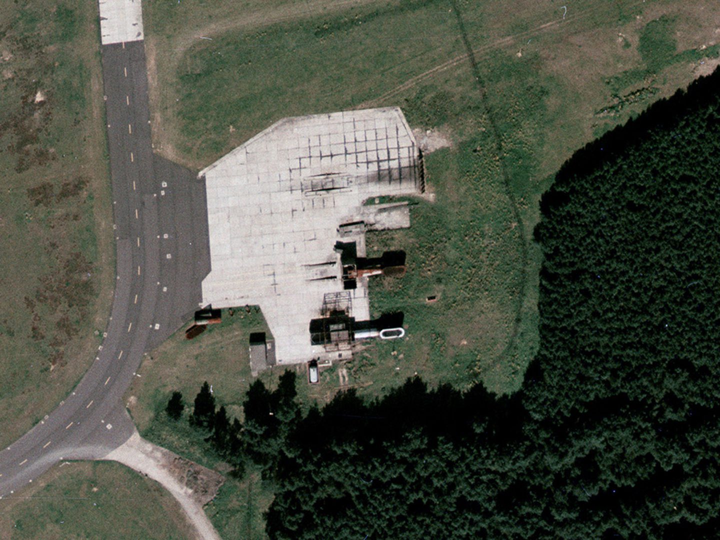 A historic colour vertical aerial photo of 2 engine detuners at the edge of an area of light-coloured concrete, surrounded by grass and next to a wood. The detuuners are cylindrical objects on their sides.