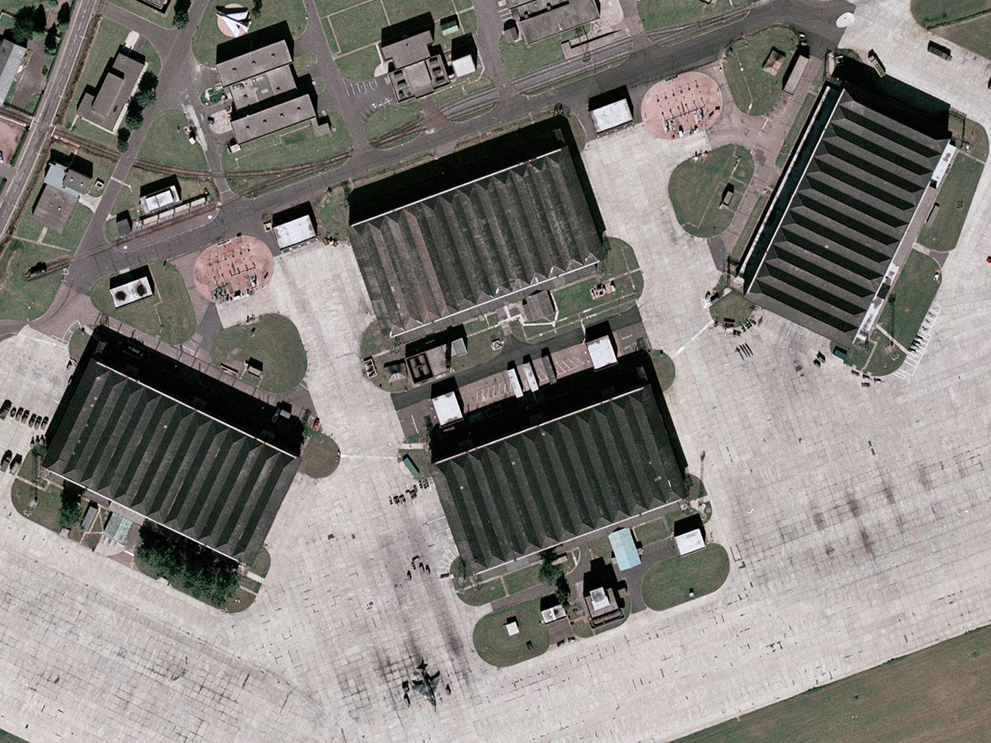 A historic colour vertical aerial photo of four large aircraft hangars with grey, sawtooth roofs. The hangars are surrounded by a light-coloured area of concrete.