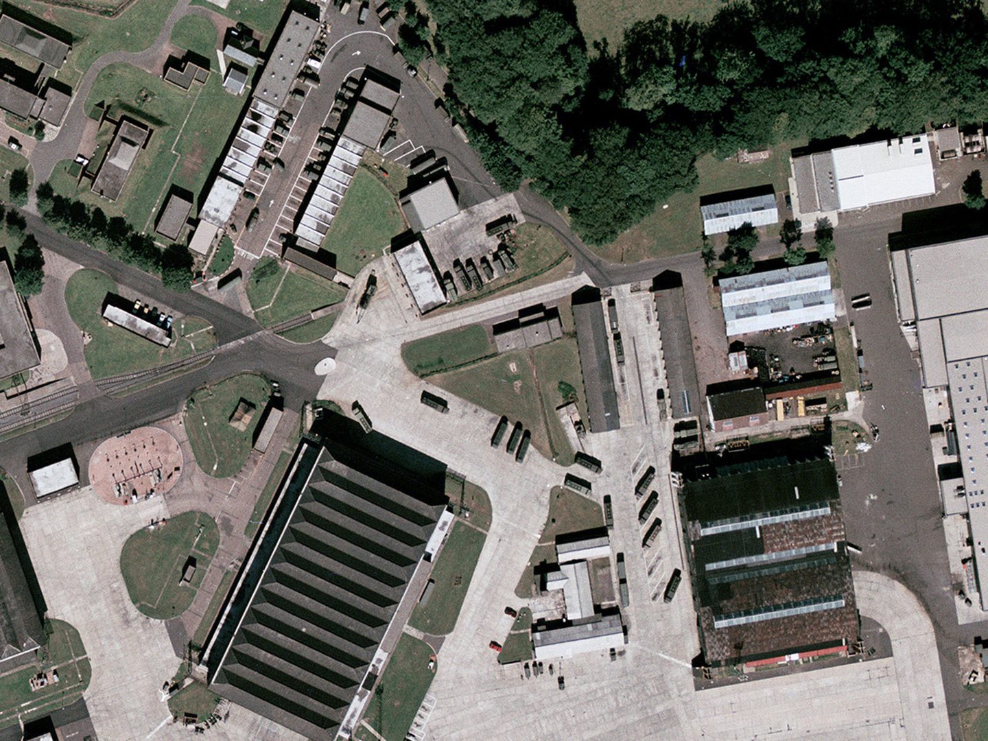 A historic colour vertical aerial photo of a collection of buildings - including a large hangar with a grey sawtooth roof - linked by roads and areas of light-coloured concrete. Numerous vehicles are parked around the buildings.