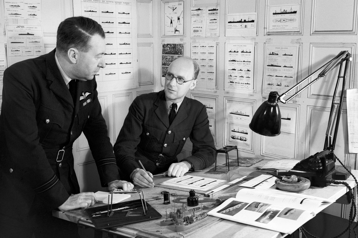 A black and white historic photo of two men in uniform at a desk. There are stereoscope viewers, a lamp and model ships on the desk. The walls behind the men are covered in silhouettes of numerous naval vessels.