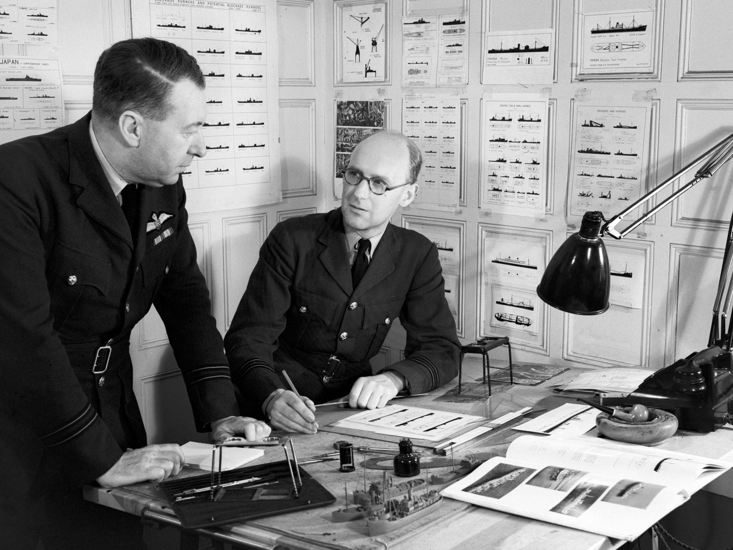 A black and white historic photo of two men in uniform at a desk. There are stereoscope viewers, a lamp and model ships on the desk. The walls behind the men are covered in silhouettes of numerous naval vessels.