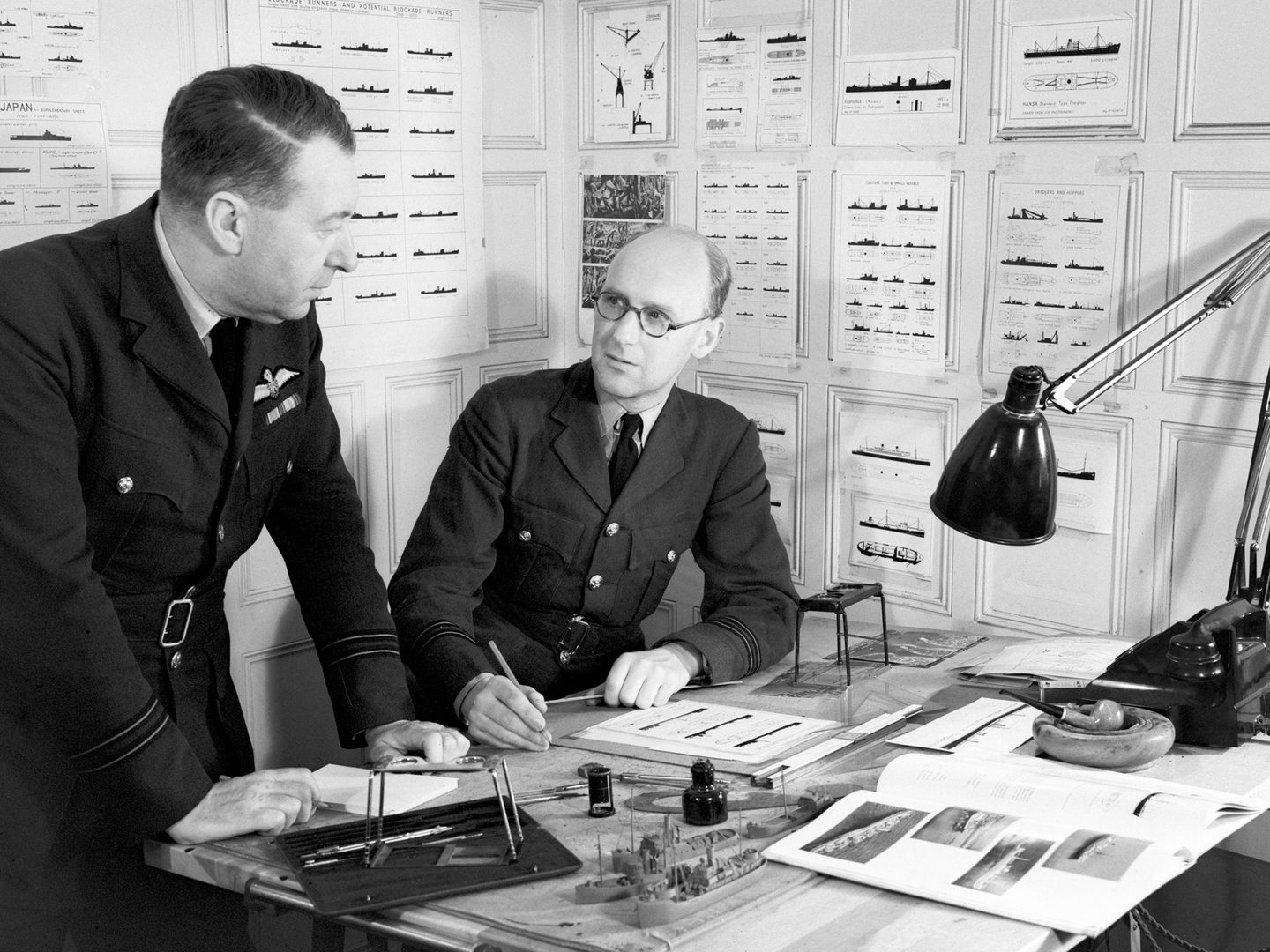 A black and white historic photo of two men in uniform at a desk. There are stereoscope viewers, a lamp and model ships on the desk. The walls behind the men are covered in silhouettes of numerous naval vessels.