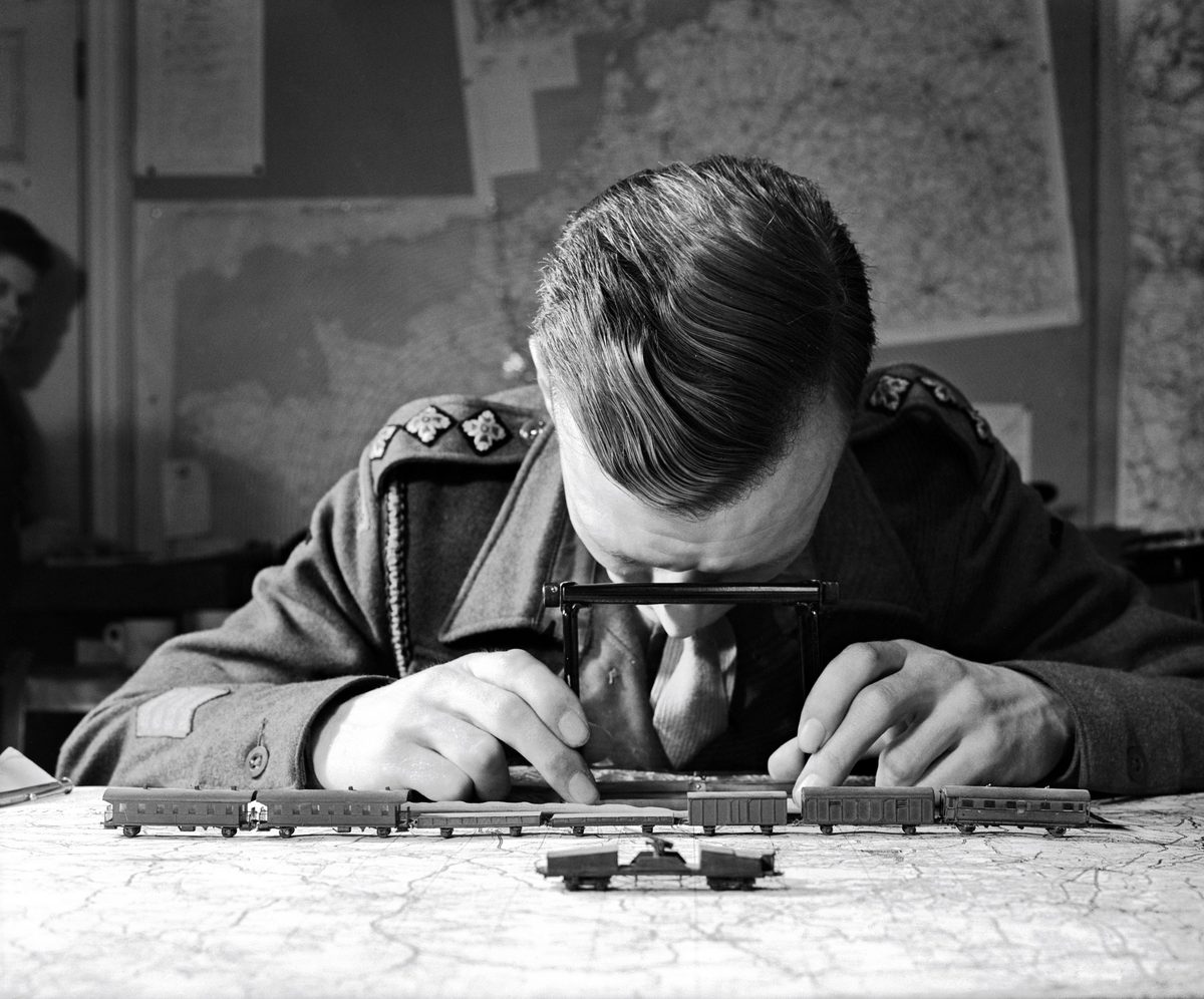 A historic black and white photo of a man in uniform sitting at a desk. On the desk in front of him is a map and a model of train carriages. He is looking down at the surface of the desk through a small stereoscope.