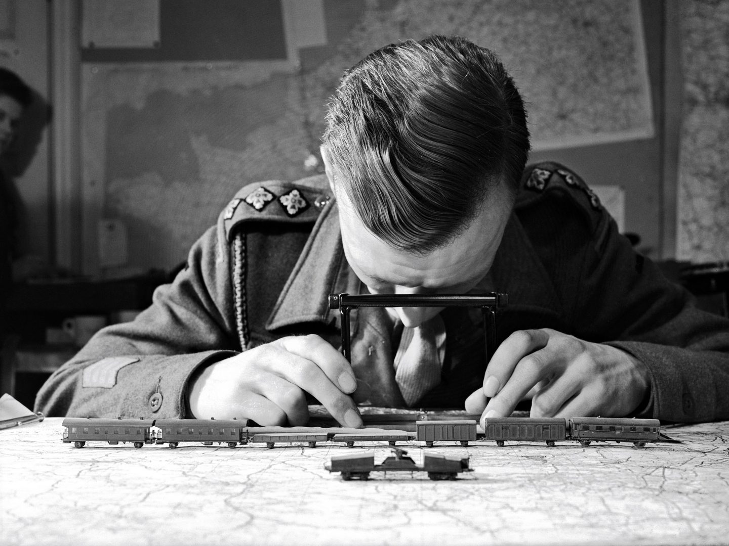 A historic black and white photo of a man in uniform sitting at a desk. On the desk in front of him is a map and a model of train carriages. He is looking down at the surface of the desk through a small stereoscope.