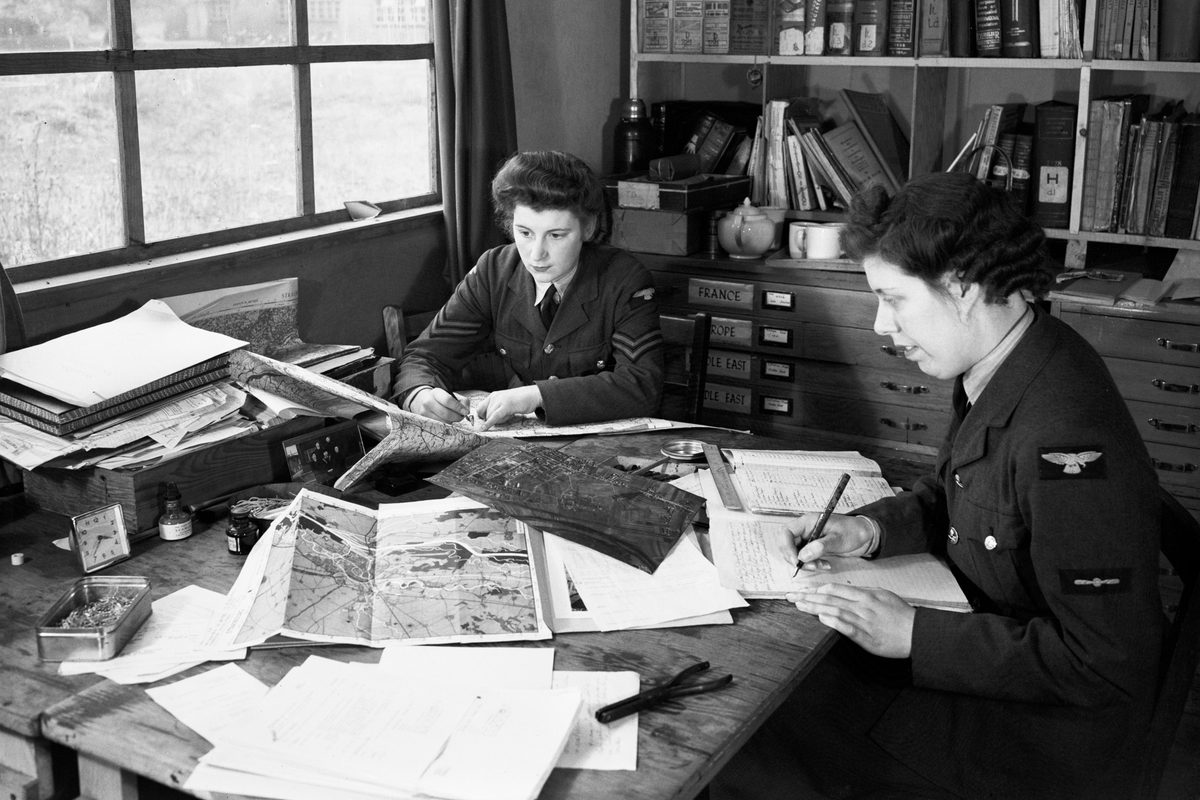 A black and white historic photo of 2 uniformed women sitting at a desk underneath a window. There are maps and aerial photos on the desk, and books on a bookshelf behind them.