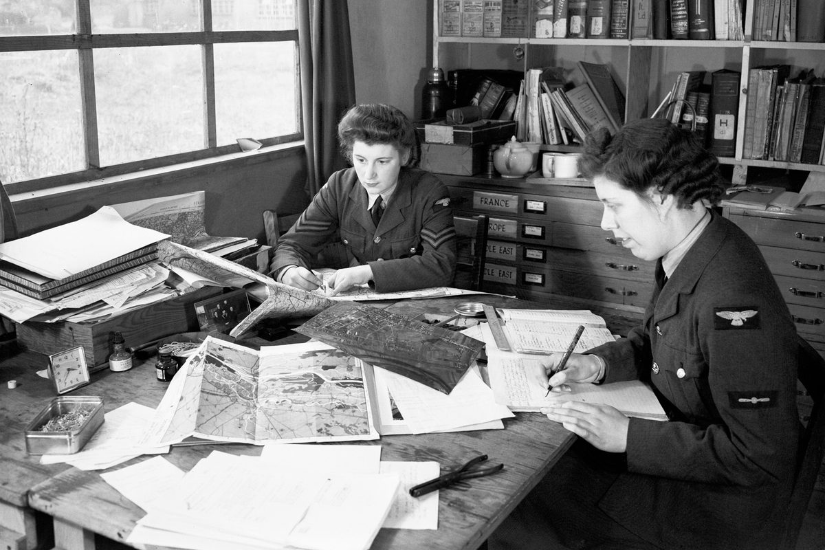 A black and white historic photo of 2 uniformed women sitting at a desk underneath a window. There are maps and aerial photos on the desk, and books on a bookshelf behind them.