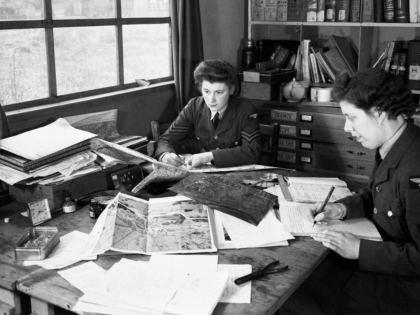 A black and white historic photo of 2 uniformed women sitting at a desk underneath a window. There are maps and aerial photos on the desk, and books on a bookshelf behind them.