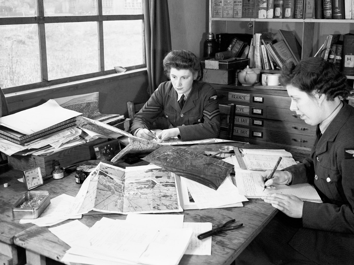 A black and white historic photo of 2 uniformed women sitting at a desk underneath a window. There are maps and aerial photos on the desk, and books on a bookshelf behind them.