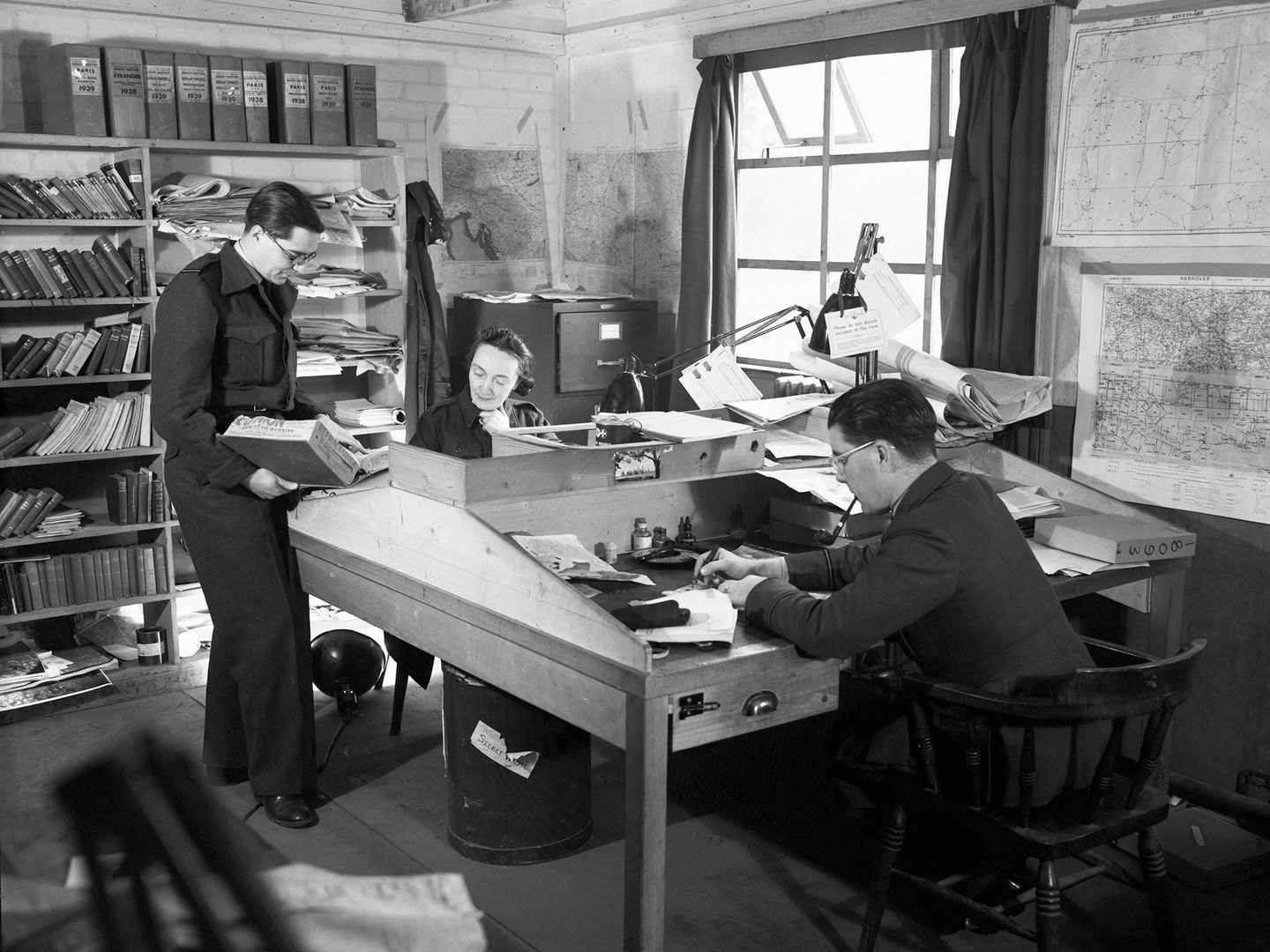 A black and white historic photo of a man and a woman sitting either side of a desk. The woman is facing us, and being shown something be a 2nd man standing to the left.