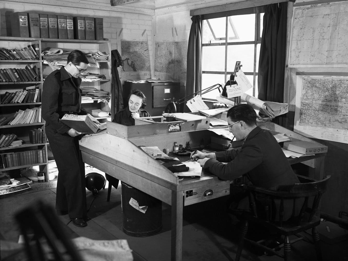 A black and white historic photo of a man and a woman sitting either side of a desk. The woman is facing us, and being shown something be a 2nd man standing to the left.
