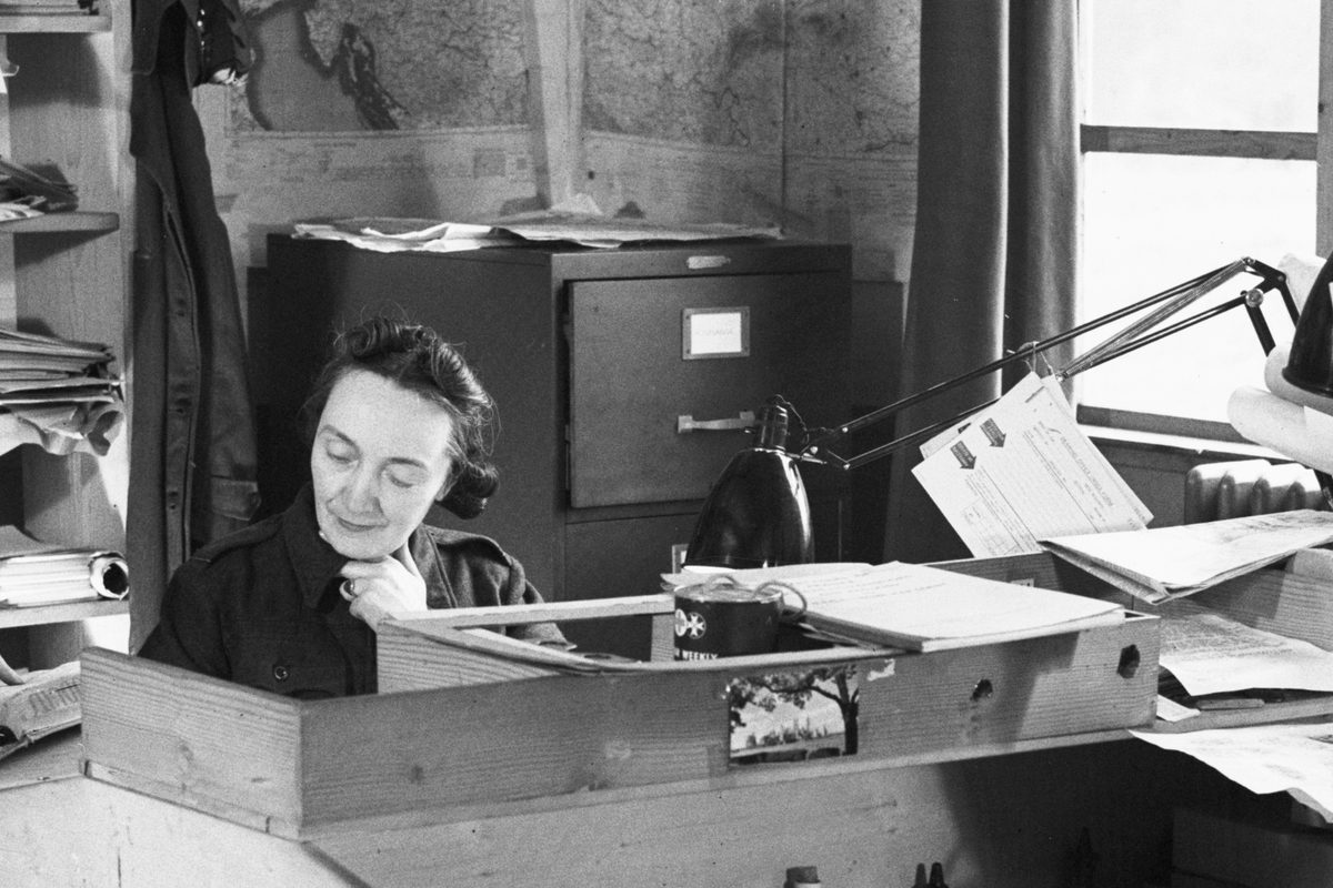 A black and white historic photo of a woman sitting at a desk. A filing cabinet and shelves are visible behind her, along with maps on the walls. To the right are a window and a lamp.