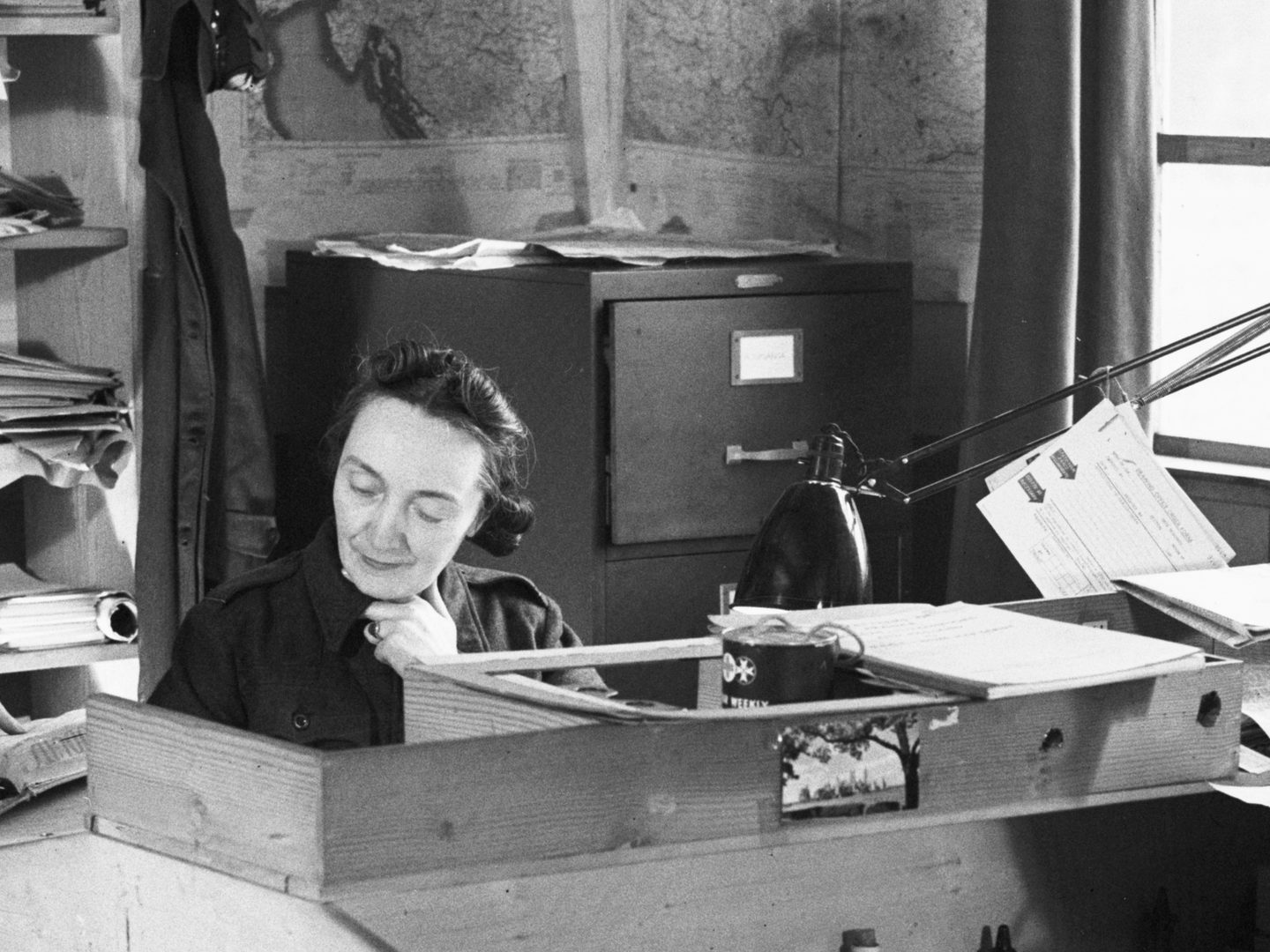 A black and white historic photo of a woman sitting at a desk. A filing cabinet and shelves are visible behind her, along with maps on the walls. To the right are a window and a lamp.