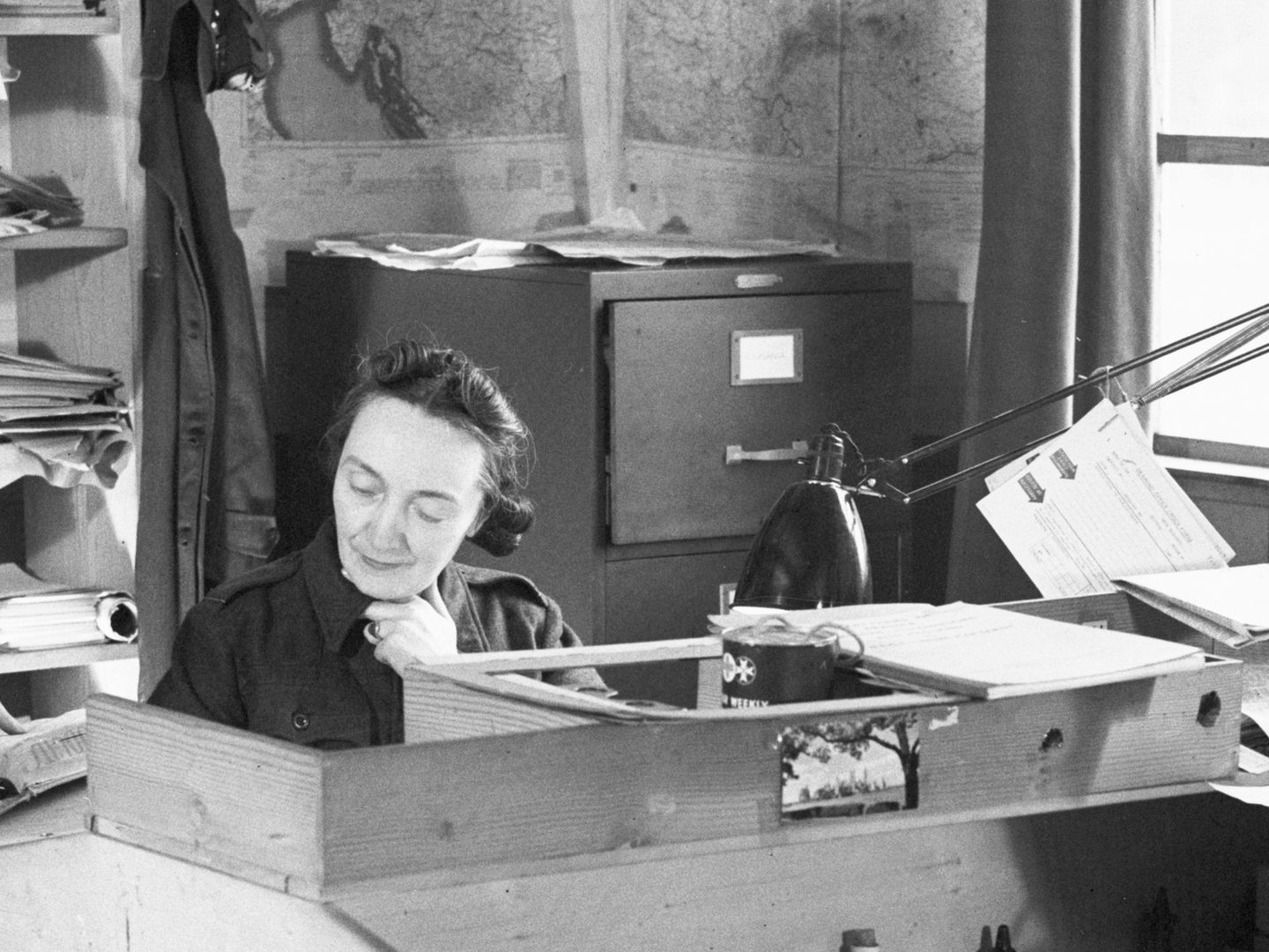 A black and white historic photo of a woman sitting at a desk. A filing cabinet and shelves are visible behind her, along with maps on the walls. To the right are a window and a lamp.