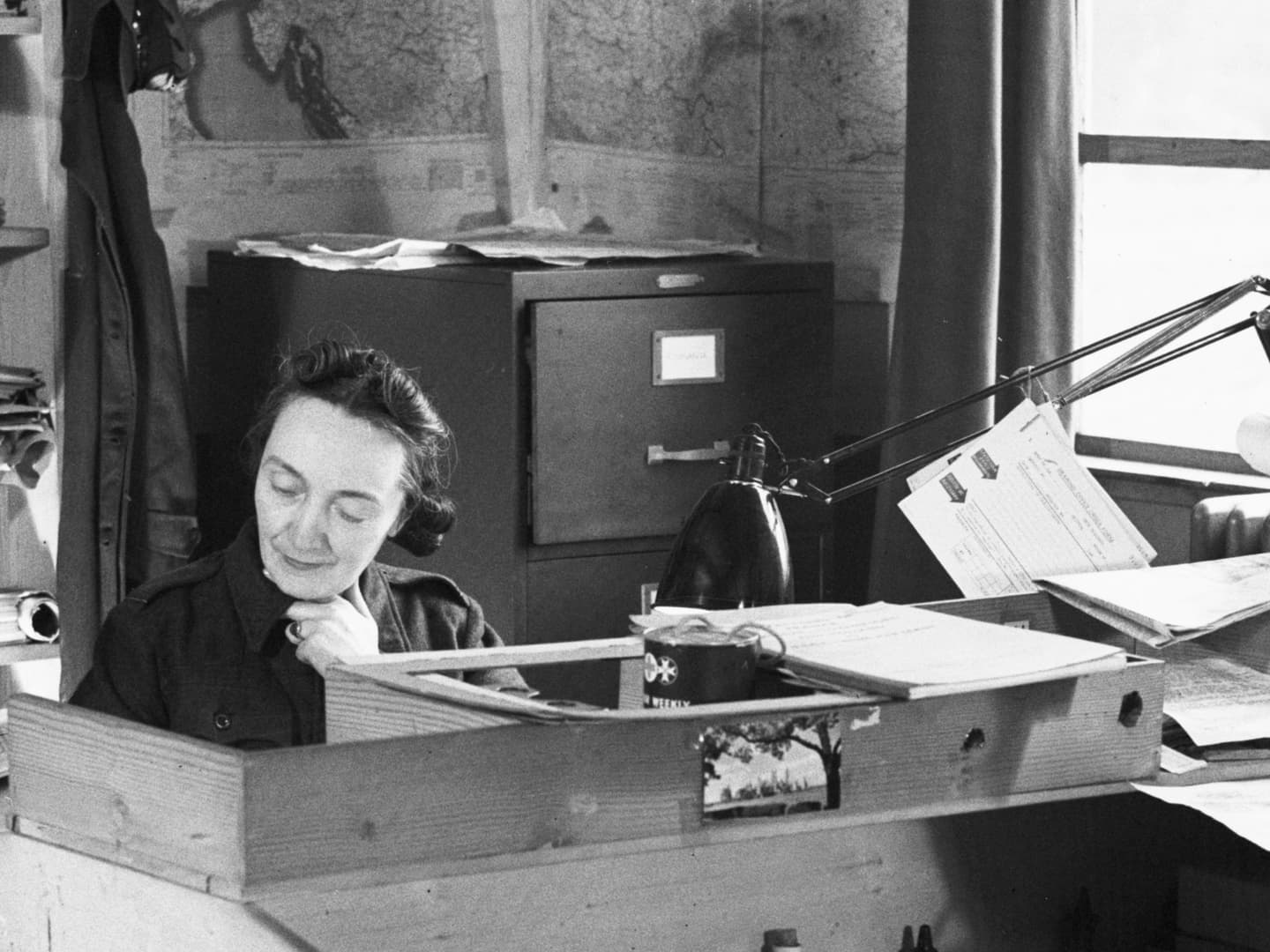 A black and white historic photo of a woman sitting at a desk. A filing cabinet and shelves are visible behind her, along with maps on the walls. To the right are a window and a lamp.