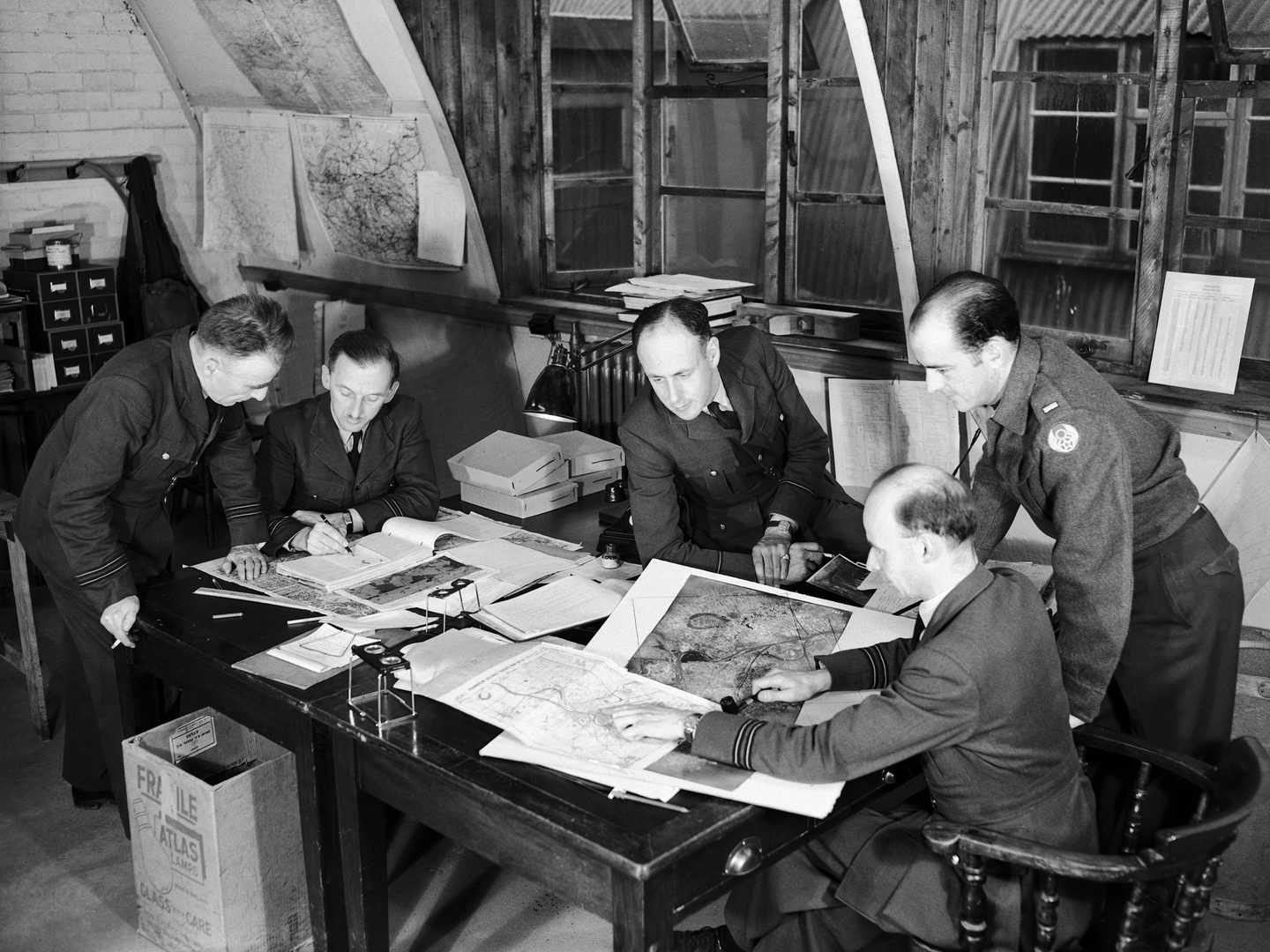 A black and white historic photo of 5 men in uniform around a table. They are studying various photos and maps. 2 pocket stereoscopes are also visible. 2 of the men are seated, 2 standing and 1 leans on the table. 3 of them are smoking.