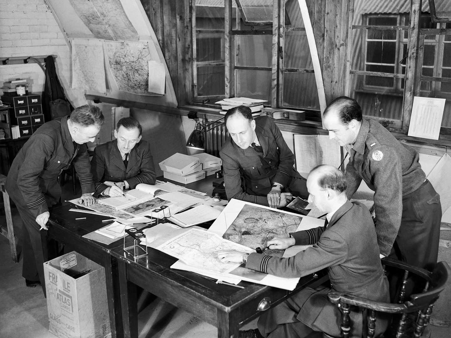 A black and white historic photo of 5 men in uniform around a table. They are studying various photos and maps. 2 pocket stereoscopes are also visible. 2 of the men are seated, 2 standing and 1 leans on the table. 3 of them are smoking.