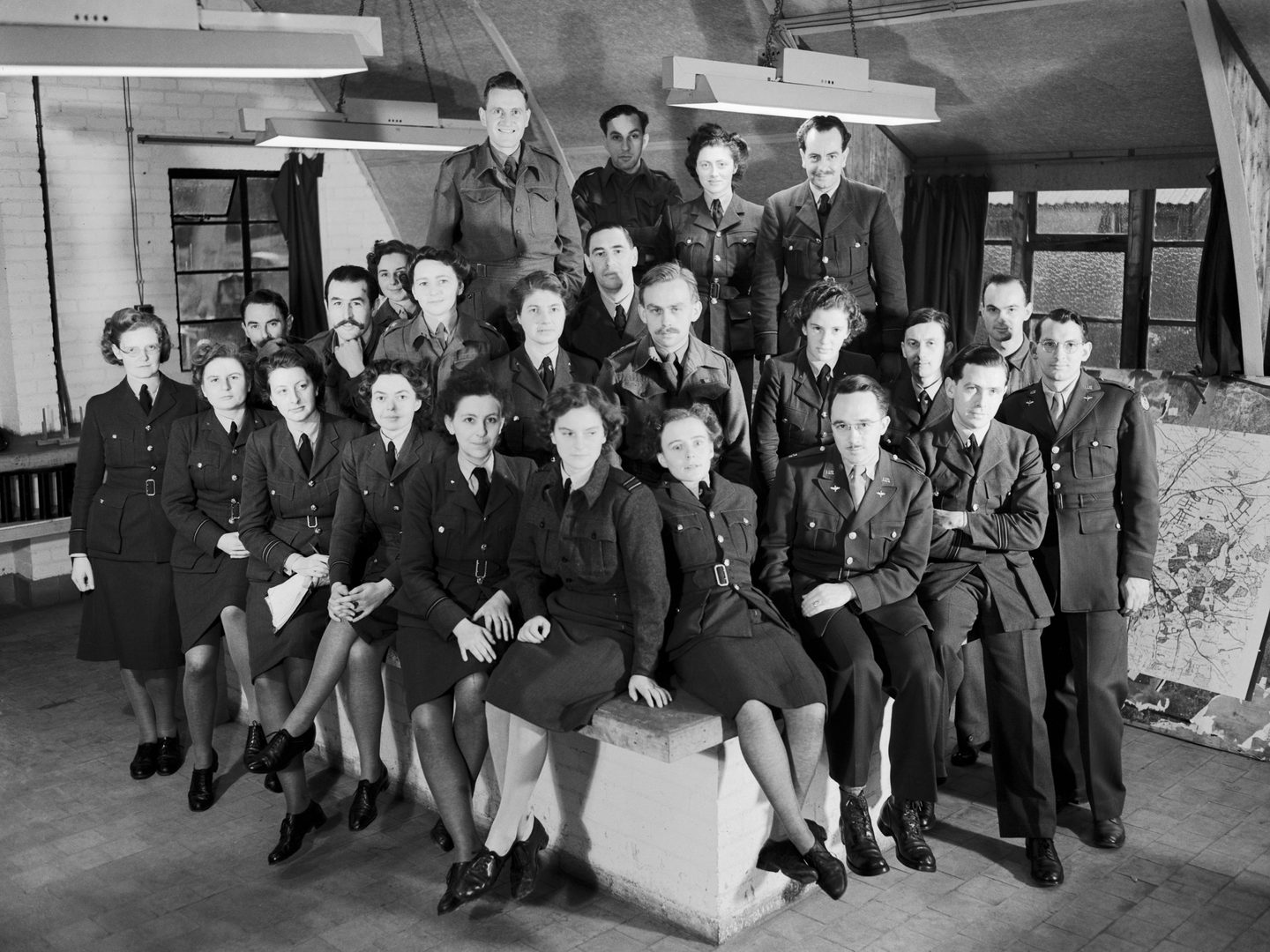A black and white historic photo of a group of about 20 uniformed women and men in an interior, some sitting and some standing, all looking at the camera.
