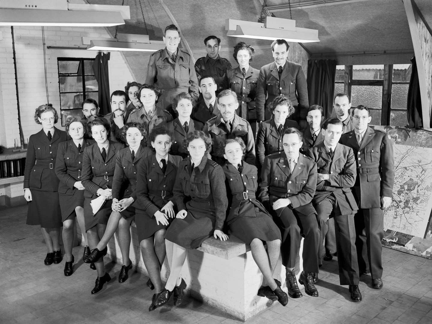 A black and white historic photo of a group of about 20 uniformed women and men in an interior, some sitting and some standing, all looking at the camera.