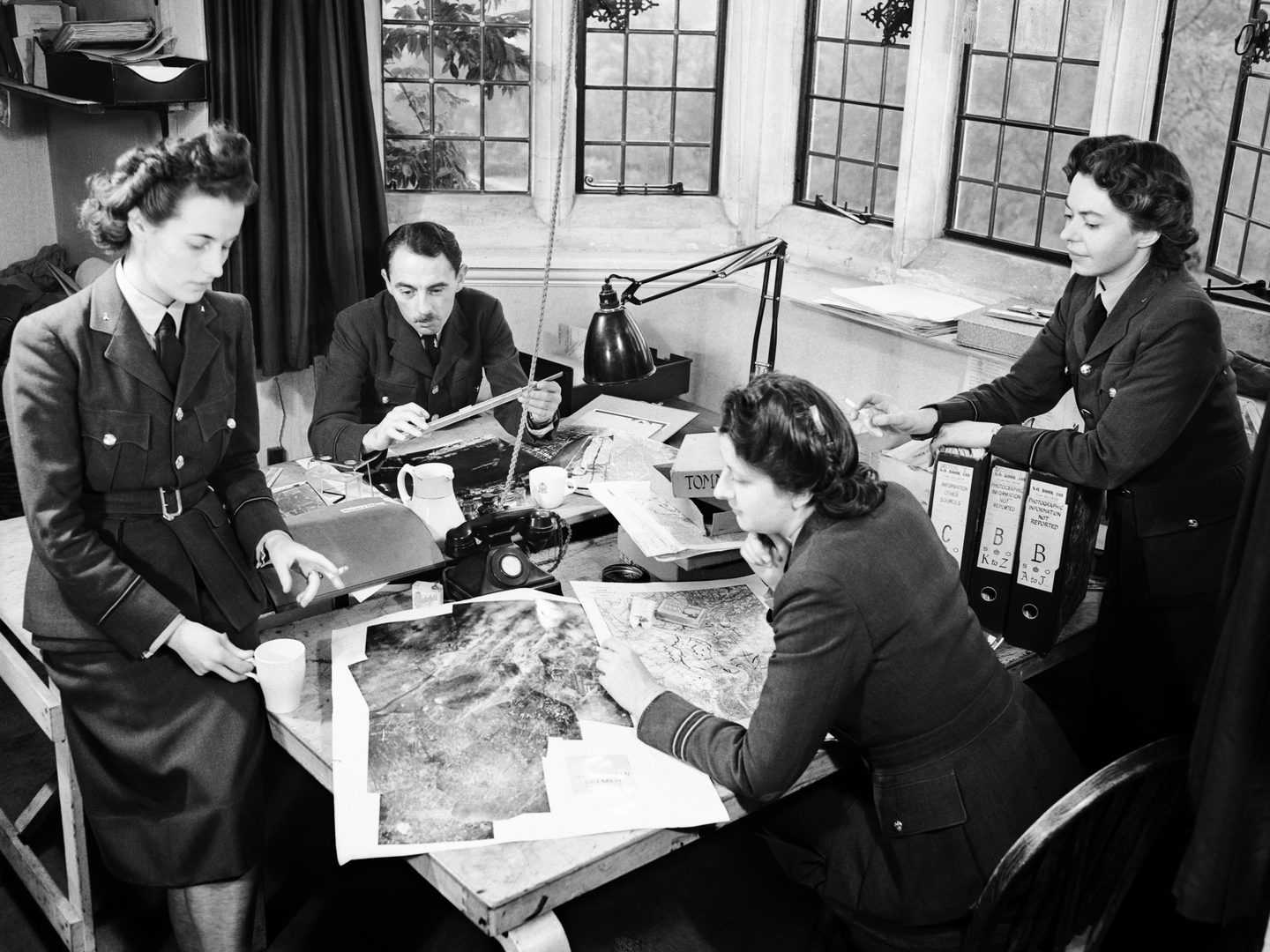 A black and white historic photo of 4 uniformed people around a table. 2 women stand to the left and right, while a 3rd woman sits on a chair in front of the table and a man sits behind it. A lamp and mosaic of aerial photos are on the table.