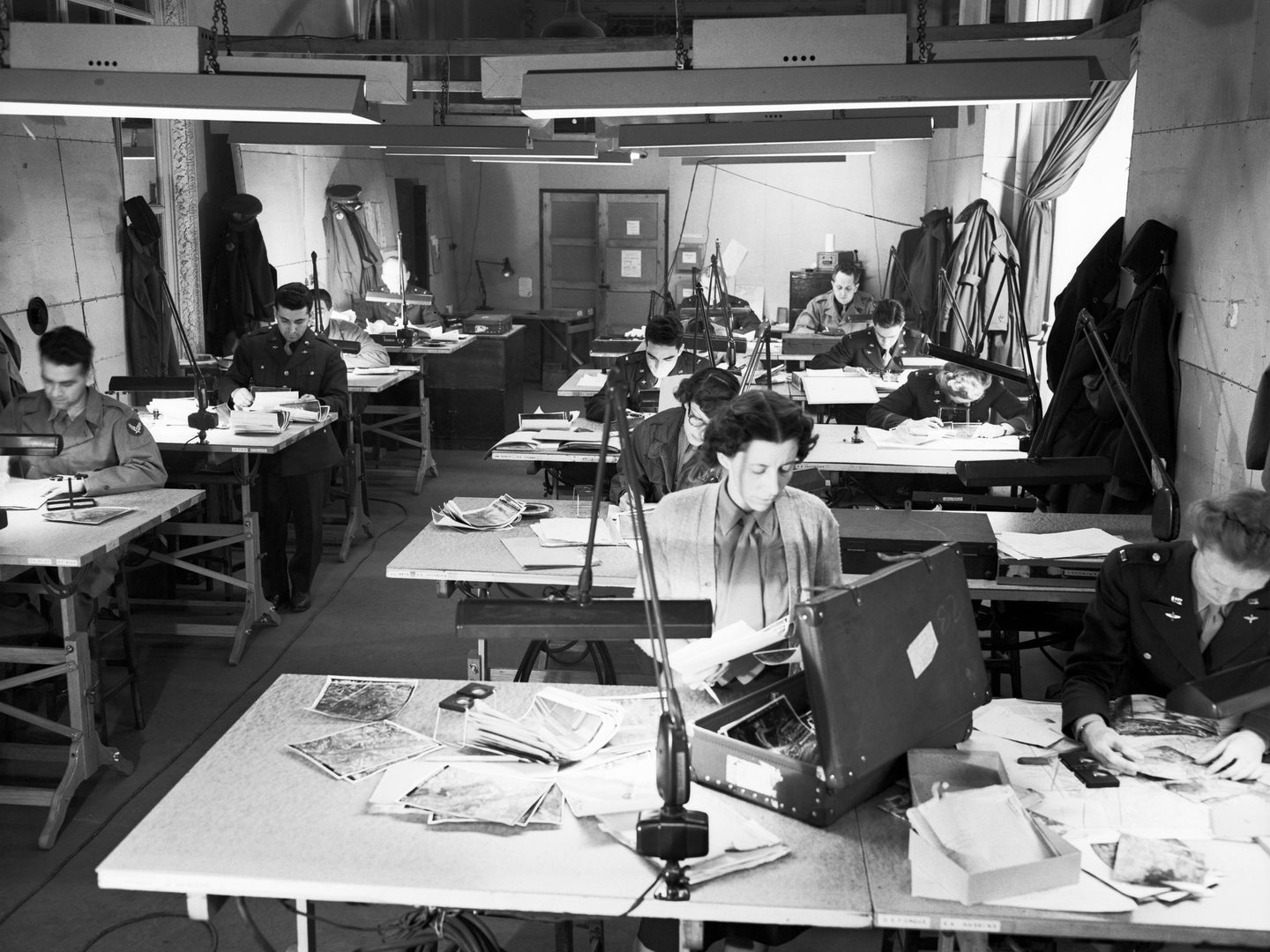 A black and white historic photo of a room full of desks. Men and women in uniform sit at the desks, which are covered in photographic prints. Great coats hang on the walls and strip lights hang from the low ceiling.