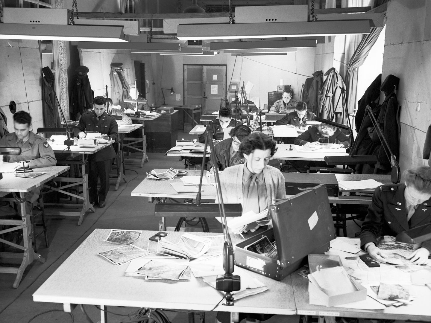 A black and white historic photo of a room full of desks. Men and women in uniform sit at the desks, which are covered in photographic prints. Great coats hang on the walls and strip lights hang from the low ceiling.