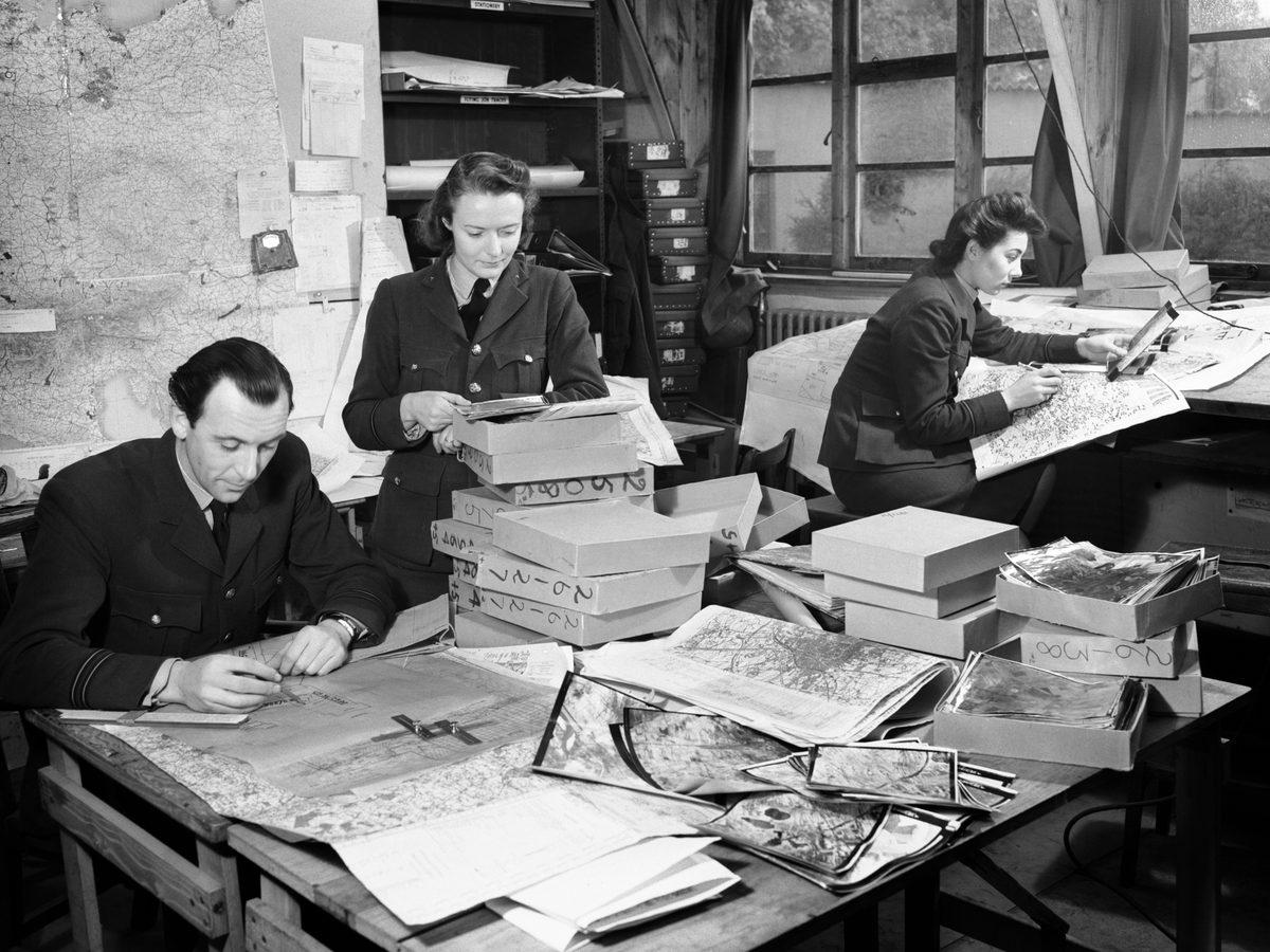 A black and white photo of a man and woman in uniform leaning over a desk. The woman stands with the man seated to her right. The desk is covered with maps, boxes and aerial photos. A 2nd uniformed woman sits at a 2nd desk, comparing a map and photo.