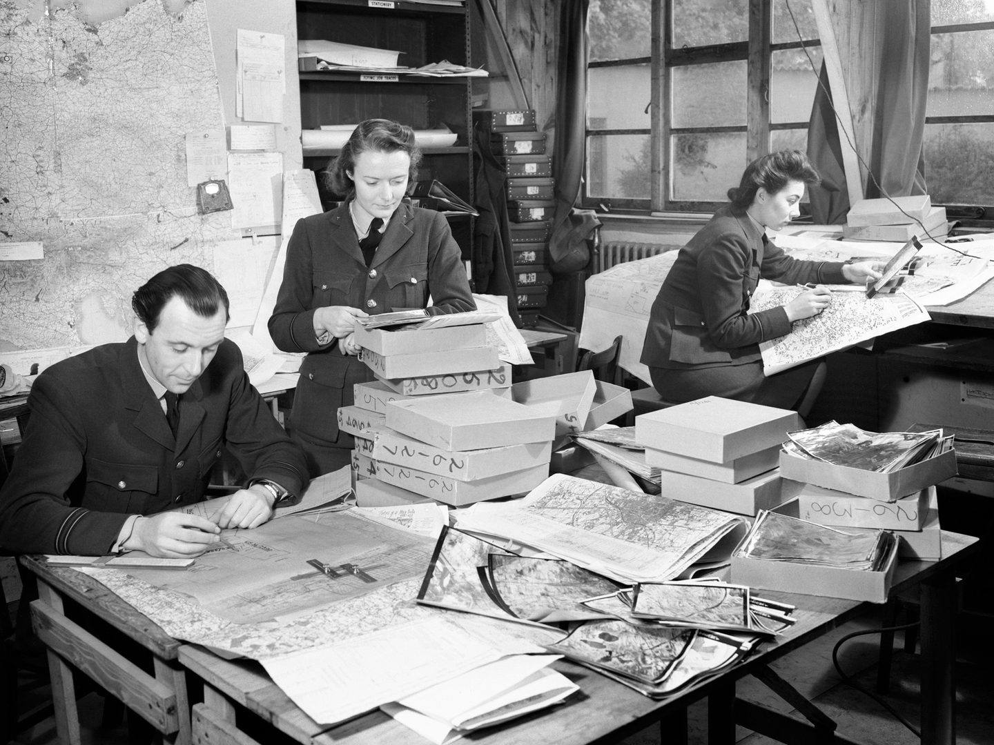 A black and white photo of a man and woman in uniform leaning over a desk. The woman stands with the man seated to her right. The desk is covered with maps, boxes and aerial photos. A 2nd uniformed woman sits at a 2nd desk, comparing a map and photo.