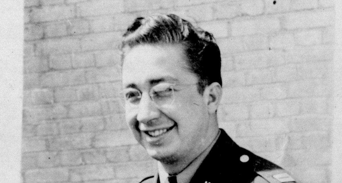 A historic black and white photo of a man wearing glasses and a uniform, standing in front of a brick wall. He is smiling, his hair is slicked back and he has a 'US' badge on his collar.