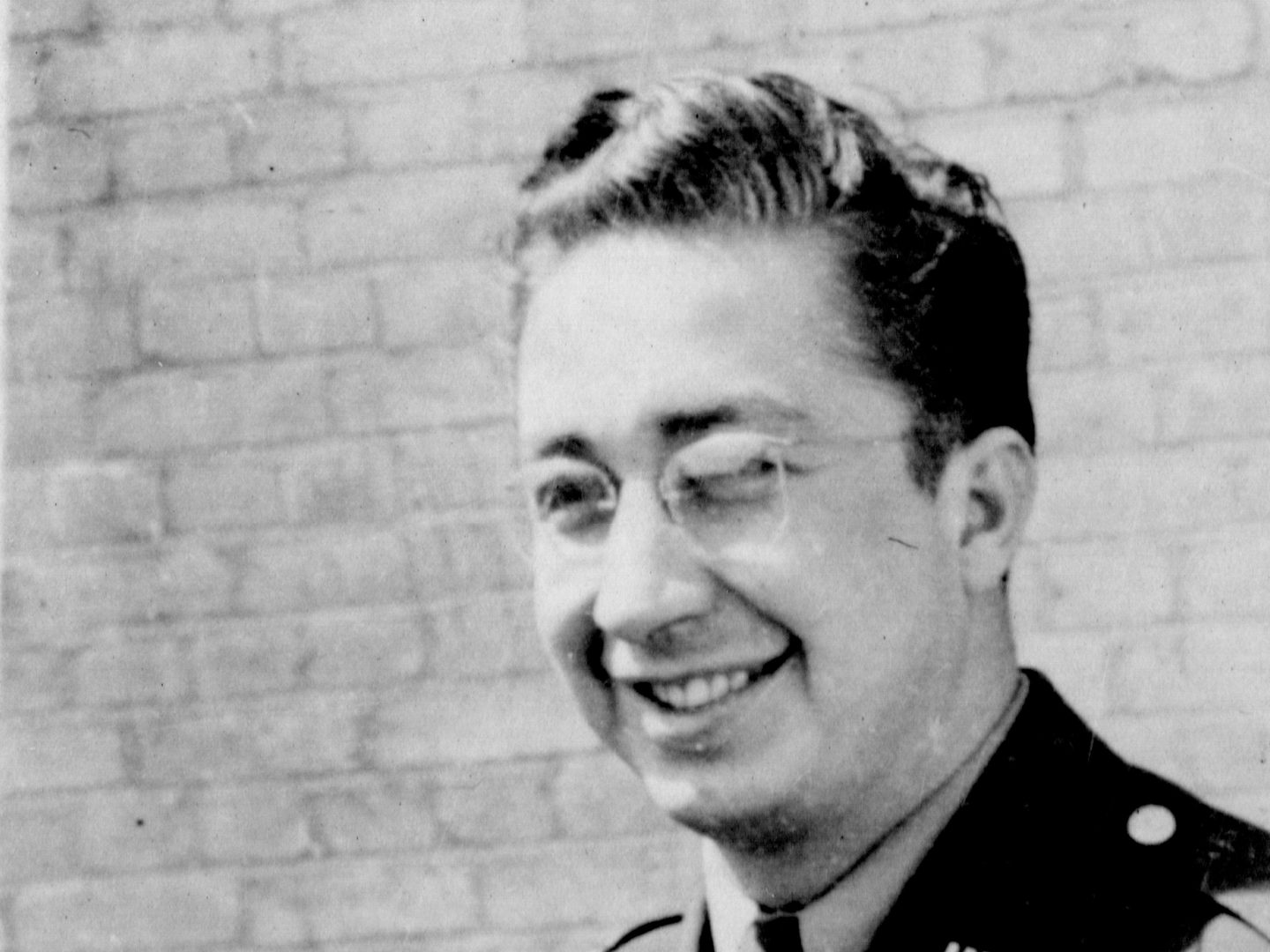 A historic black and white photo of a man wearing glasses and a uniform, standing in front of a brick wall. He is smiling, his hair is slicked back and he has a 'US' badge on his collar.
