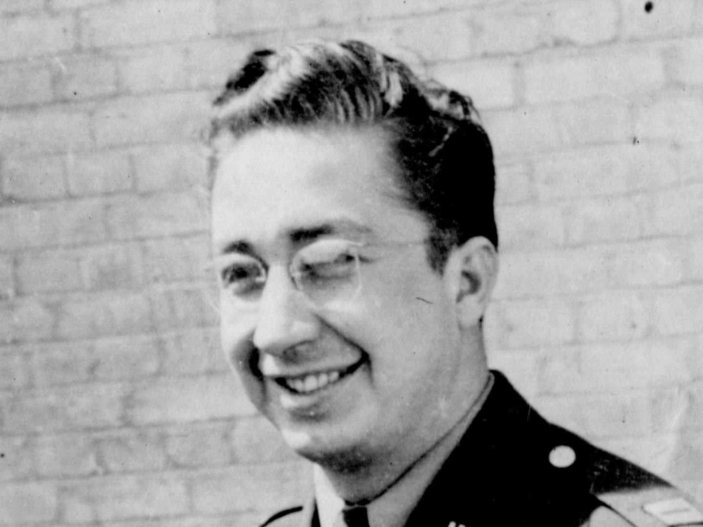 A historic black and white photo of a man wearing glasses and a uniform, standing in front of a brick wall. He is smiling, his hair is slicked back and he has a 'US' badge on his collar.