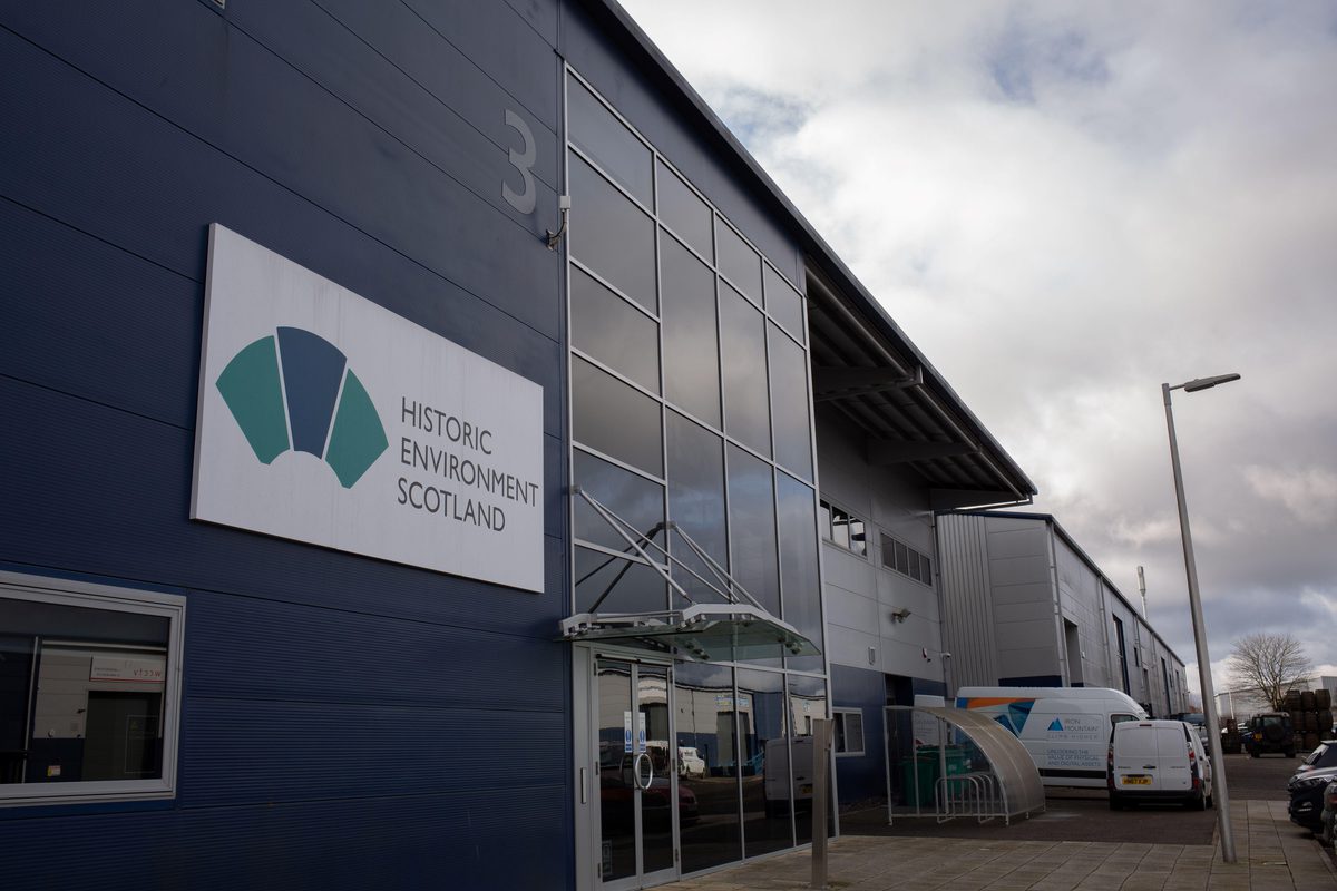 A large blue industrial building. It has the number 3 on it and the logo of Historic Environment Scotland. At the far end of the building is a bike shelter and two small white vans.