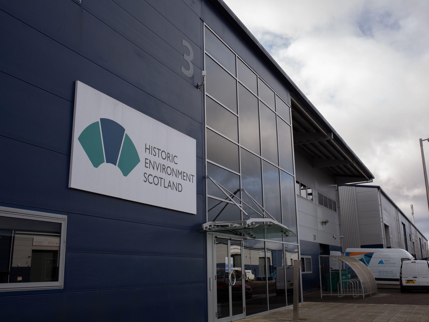 A large blue industrial building. It has the number 3 on it and the logo of Historic Environment Scotland. At the far end of the building is a bike shelter and two small white vans.