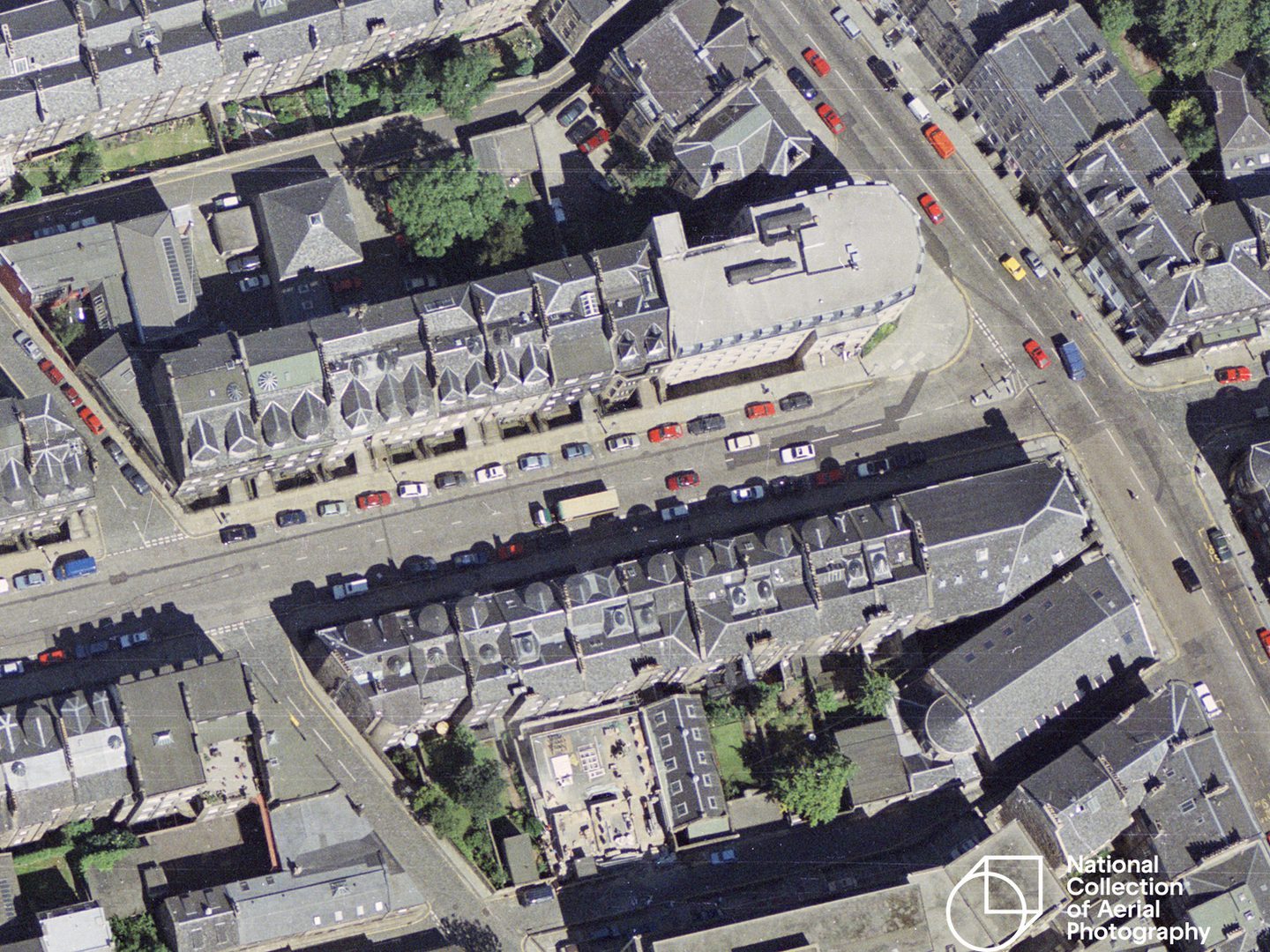 A colour aerial photo showing a grid of streets. The buildings have grey roofs and the various colours of the cars on the roads stand out, especially red.