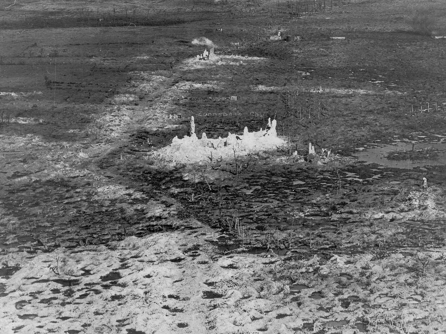 A black and white oblique aerial photo of a devastated landscape. A completely destroyed building is in the middle of the image. The ground around it is marked with shell holes and the remains of trees. There is a puff of white smoke in the distance.