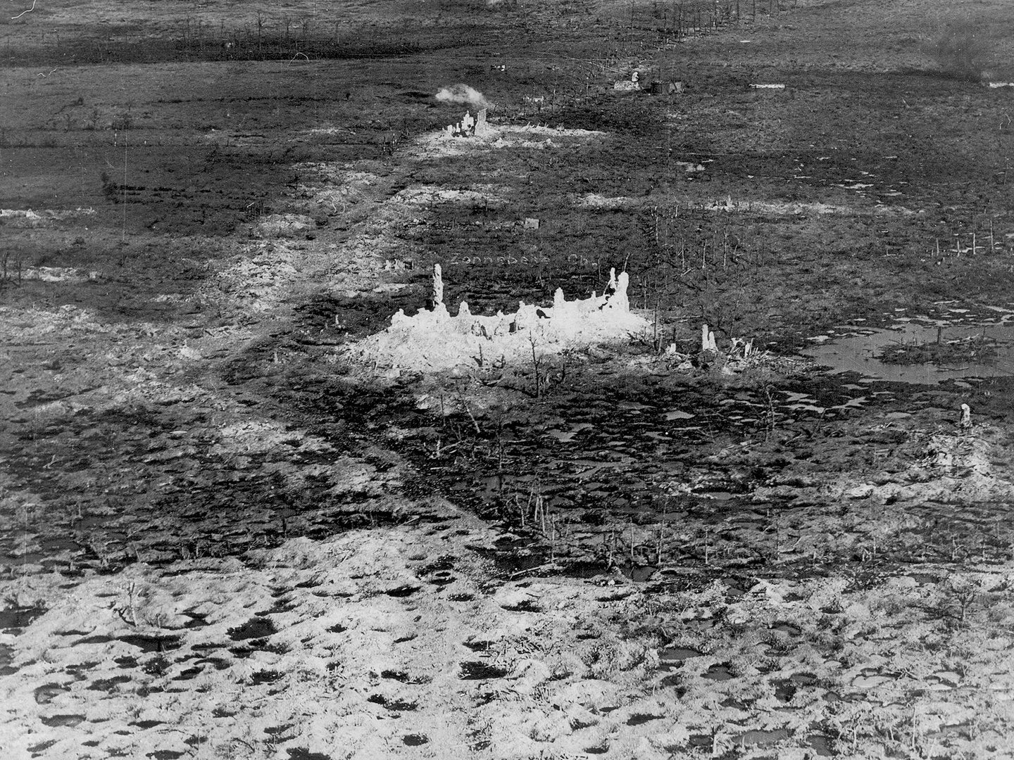 A black and white oblique aerial photo of a devastated landscape. A completely destroyed building is in the middle of the image. The ground around it is marked with shell holes and the remains of trees. There is a puff of white smoke in the distance.