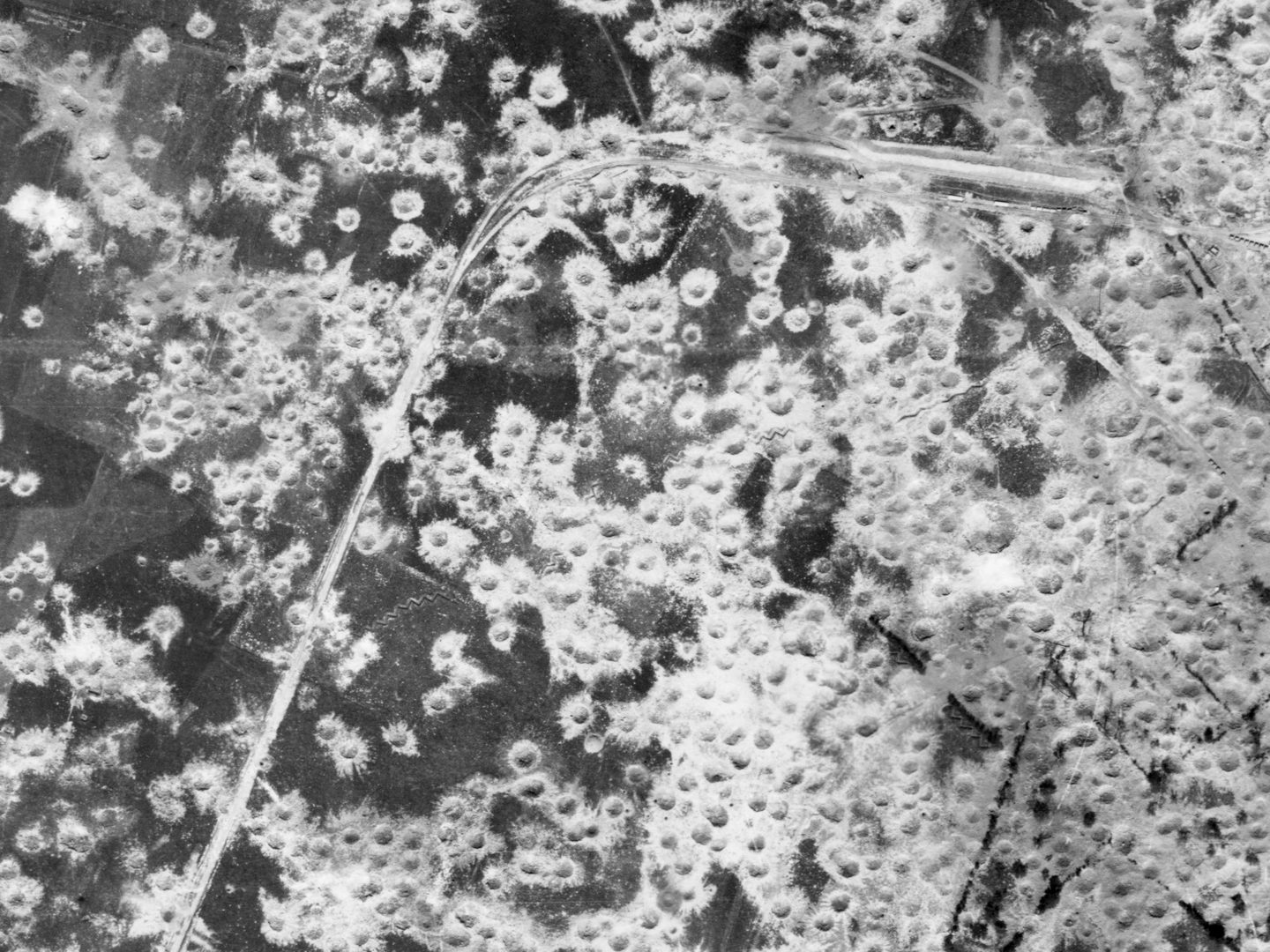A black and white aerial photo of a very heavily bombed area of land. What land is visible is dark grey, and almost obscured with numerous white bomb craters. A road can be seen running from the bottom left corner to the top right.