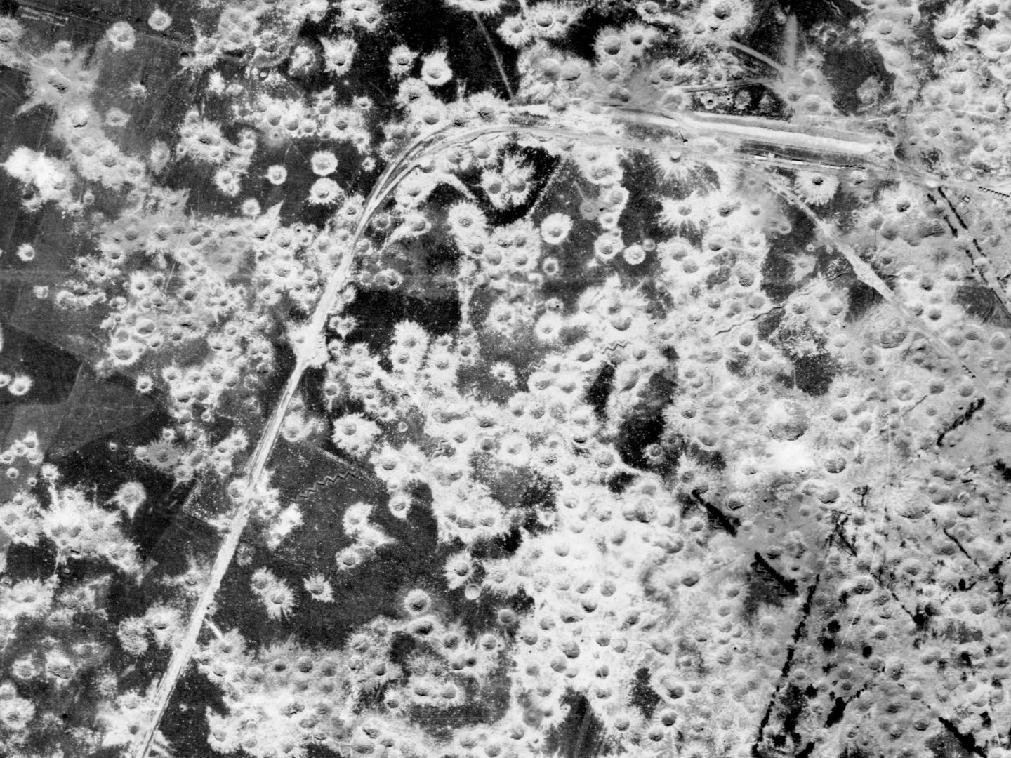 A black and white aerial photo of a very heavily bombed area of land. What land is visible is dark grey, and almost obscured with numerous white bomb craters. A road can be seen running from the bottom left corner to the top right.