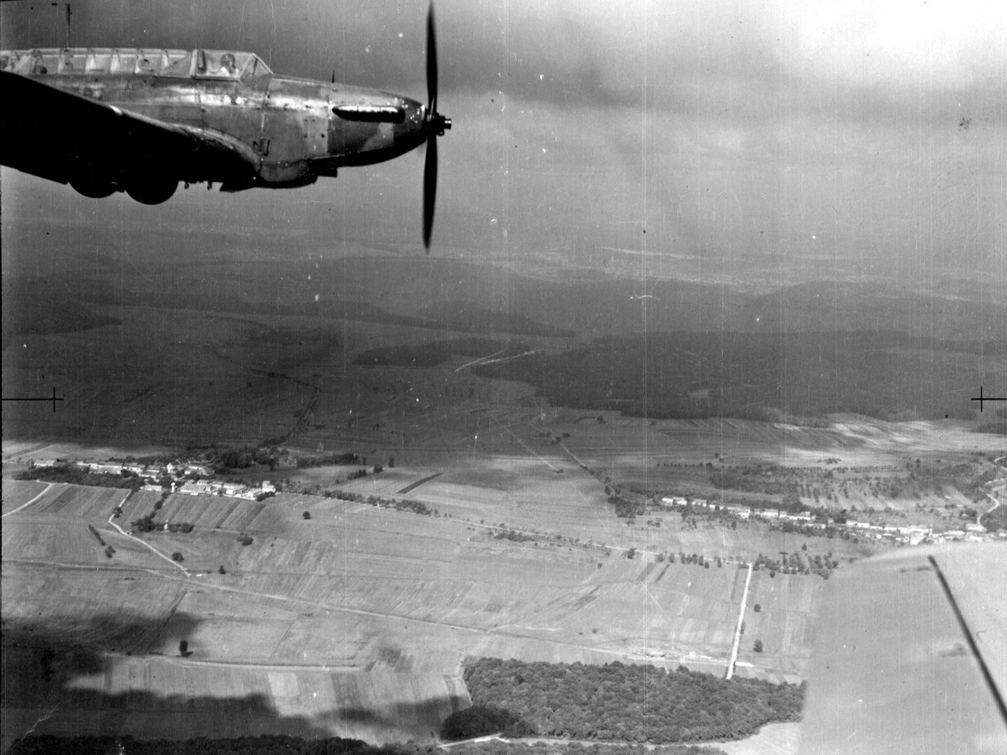 A black and white oblique aerial photo of an airborne plane – a Fairey Battle – from side-on. The plane is in the top left corner and has one propellor. It has a long, glazed cockpit with the pilot visible. Fields and woods cover the landscape below.