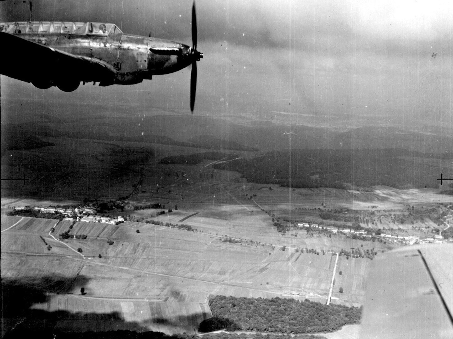A black and white oblique aerial photo of an airborne plane – a Fairey Battle – from side-on. The plane is in the top left corner and has one propellor. It has a long, glazed cockpit with the pilot visible. Fields and woods cover the landscape below.