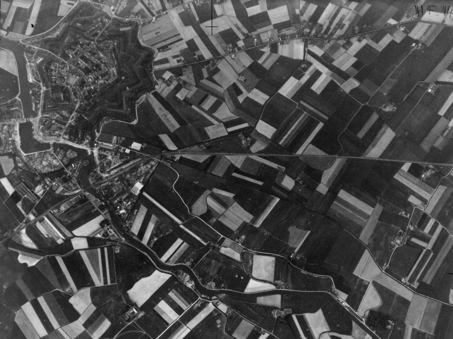A black and white aerial photo of a landscape. Most of the image is taken up with an irregular pattern of rectangular fields in various shades of grey. A fortified settlement is at the top left. A river winds along the bottom edge.
