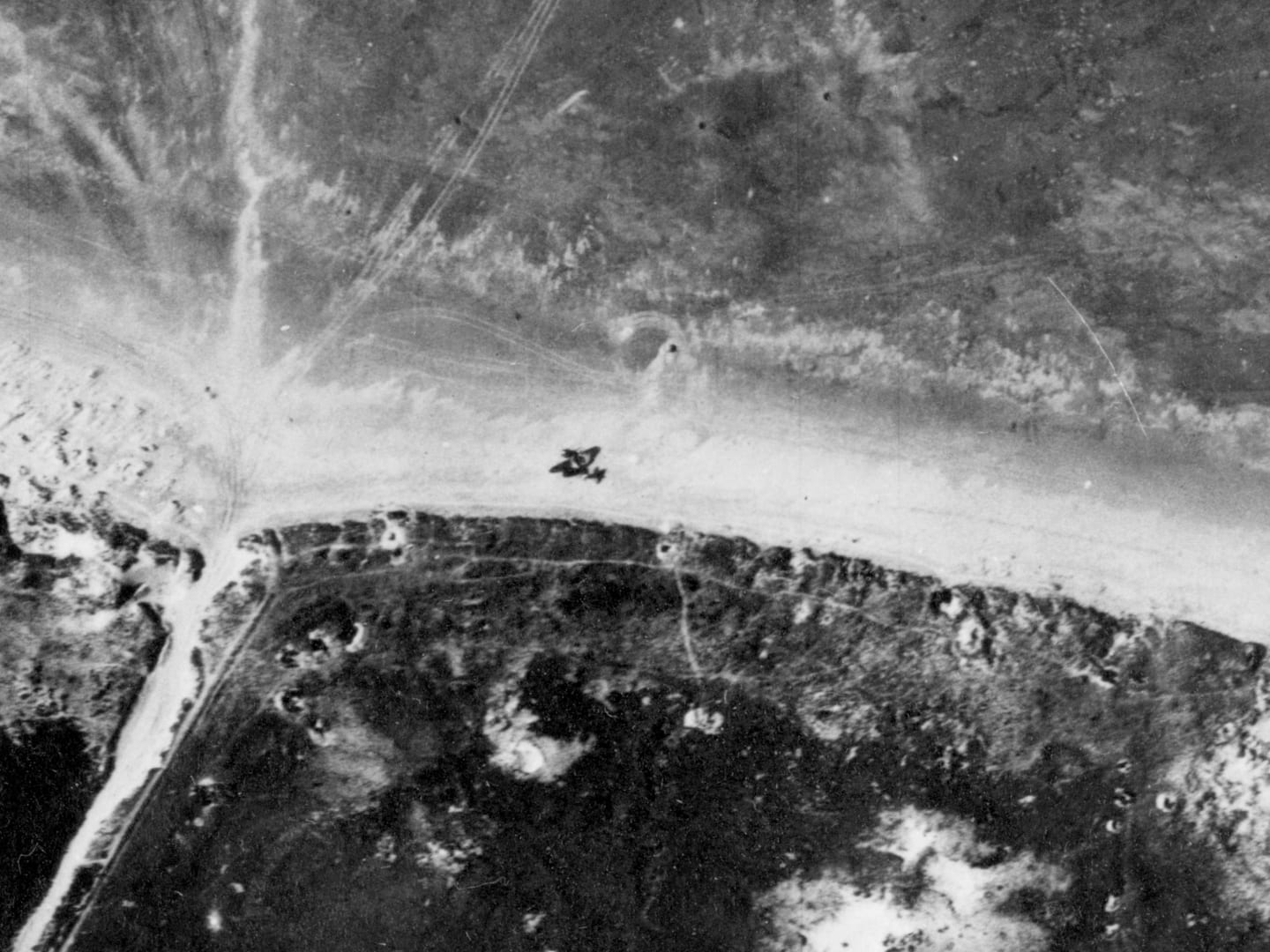 A black and white aerial photo of a sandy beach bordered by a dark shoreline. The beach is marked with craters and vehicle tracks. Lying on the beach is the black shape of a crashed plane, with its tail broken off.