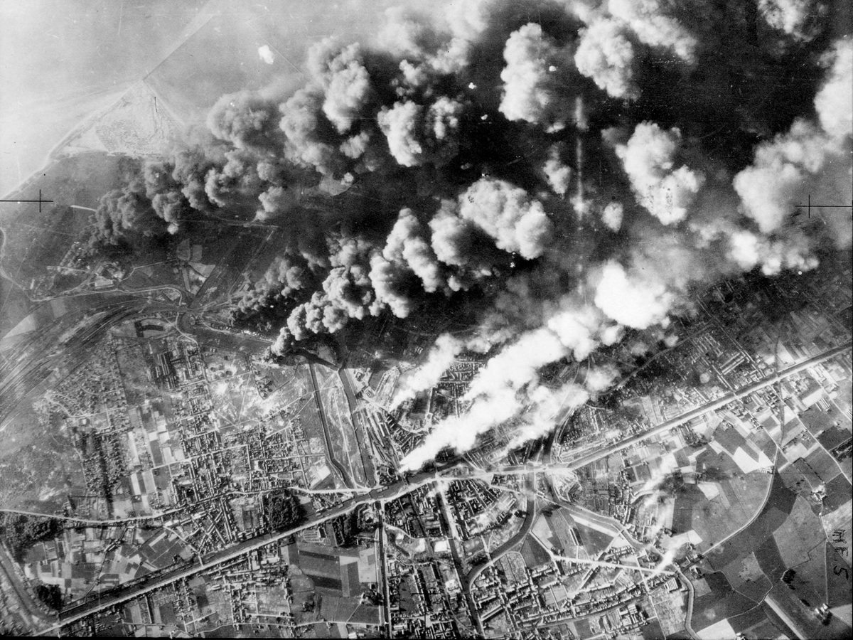 A black and white aerial photo of the city of Dunkirk, with the coast just visible in the distance. Thick clouds of black and white smoke are billowing up from the city, being driven by the wind from left to right.
