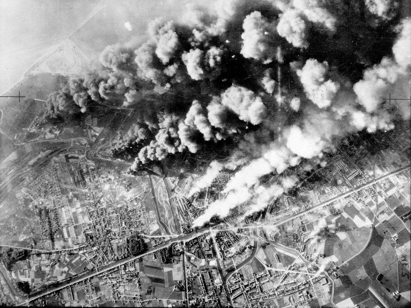 A black and white aerial photo of the city of Dunkirk, with the coast just visible in the distance. Thick clouds of black and white smoke are billowing up from the city, being driven by the wind from left to right.