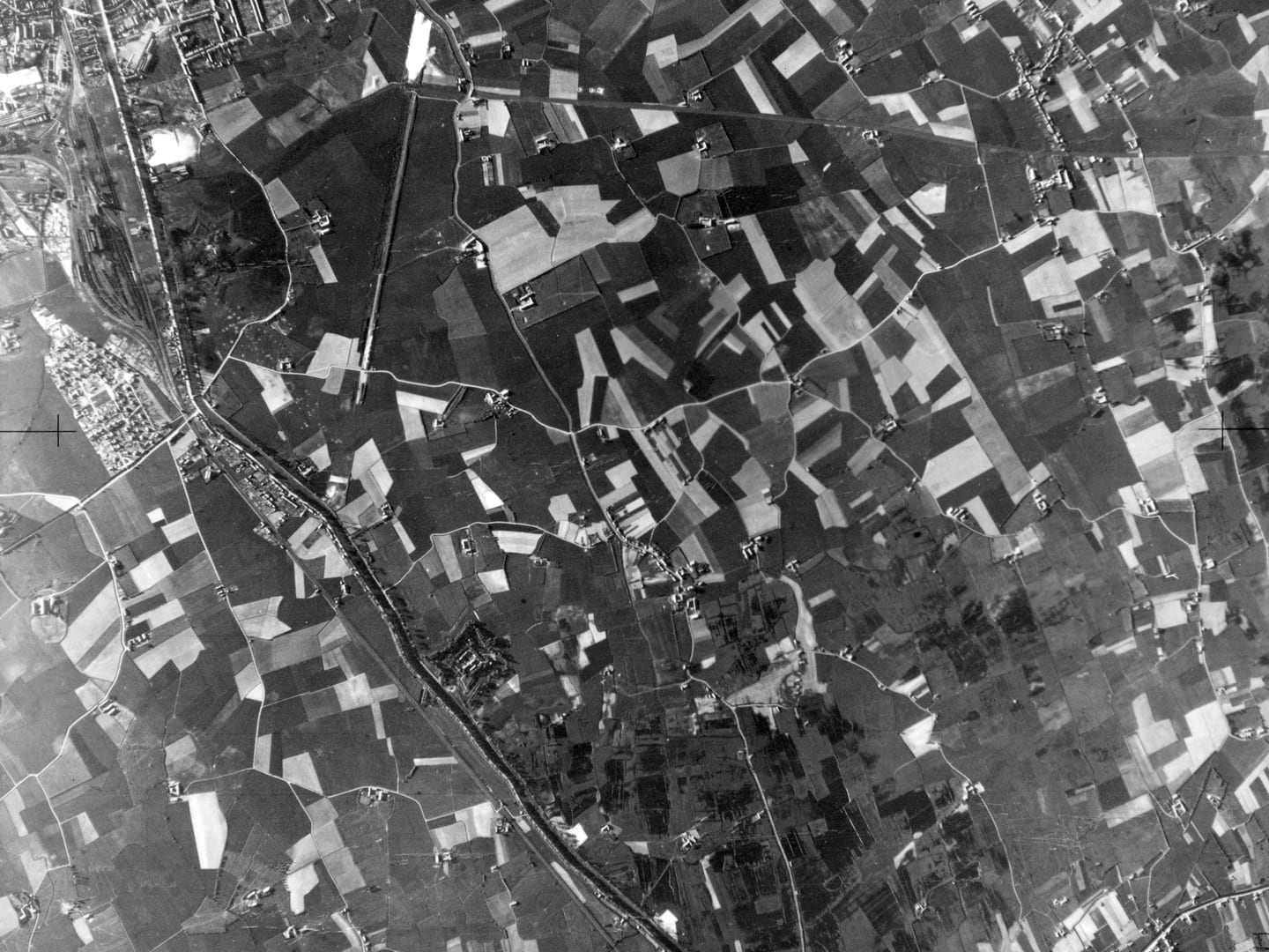 A black and white aerial photo of a patchwork of fields, crisscrossed by roads and tracks. A canal runs from the top left corner to the middle of the bottom edge. Farm buildings are scattered throughout, and there is the edge of a town top left.
