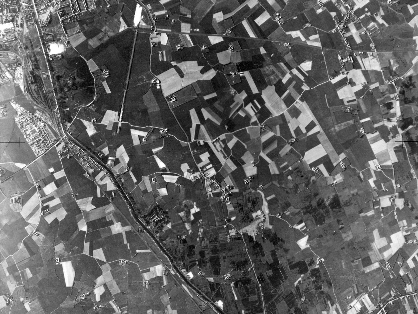 A black and white aerial photo of a patchwork of fields, crisscrossed by roads and tracks. A canal runs from the top left corner to the middle of the bottom edge. Farm buildings are scattered throughout, and there is the edge of a town top left.