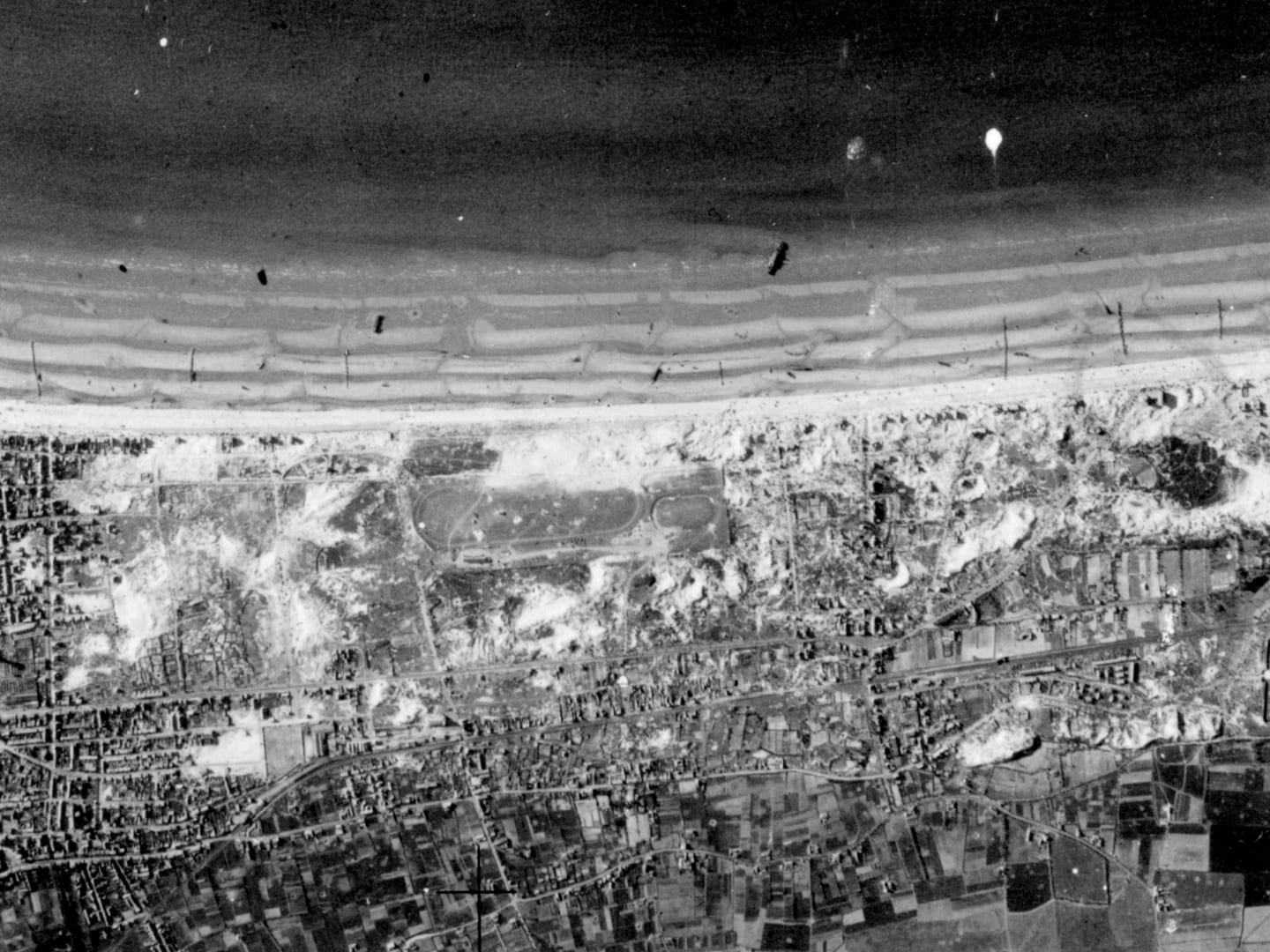 A black and white aerial photo of a sandy beach sandwiched between the sea and the shoreline. There are houses on the shoreline, which is heavily cratered and marked from bombing. There are the black shapes of boats and vehicles on the beach.