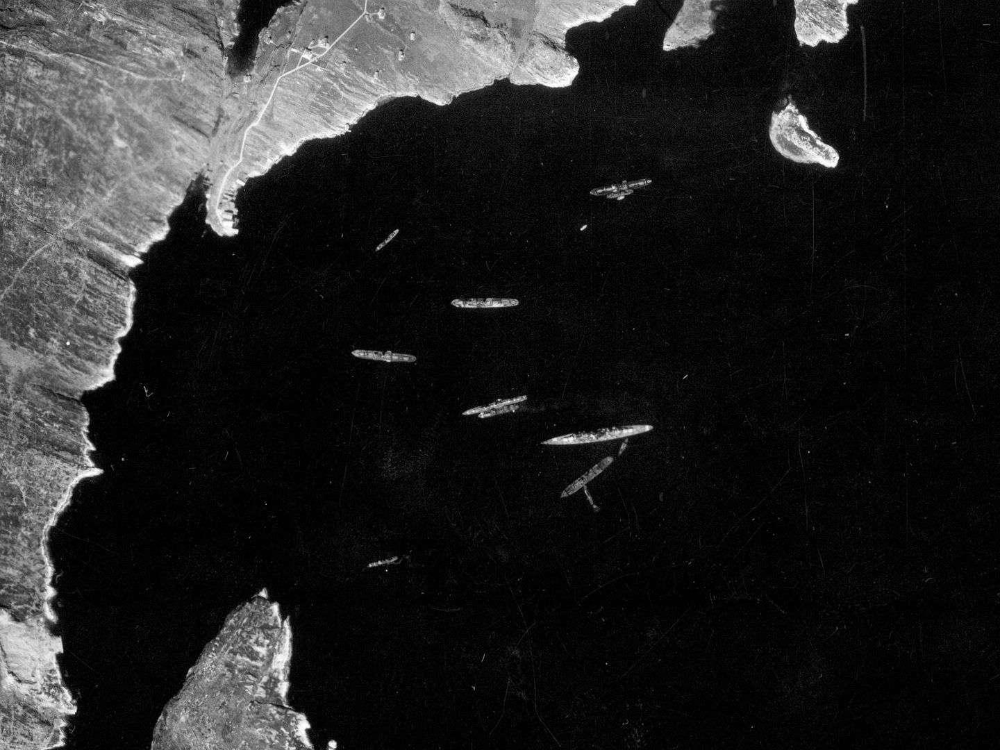 A historic black and white vertical aerial photo of a bay surrounded by a ragged coastline. In the centre of the bay is a large ship, surrounded by about 10 smaller ships.