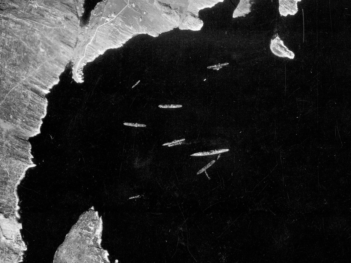 A historic black and white vertical aerial photo of a bay surrounded by a ragged coastline. In the centre of the bay is a large ship, surrounded by about 10 smaller ships.